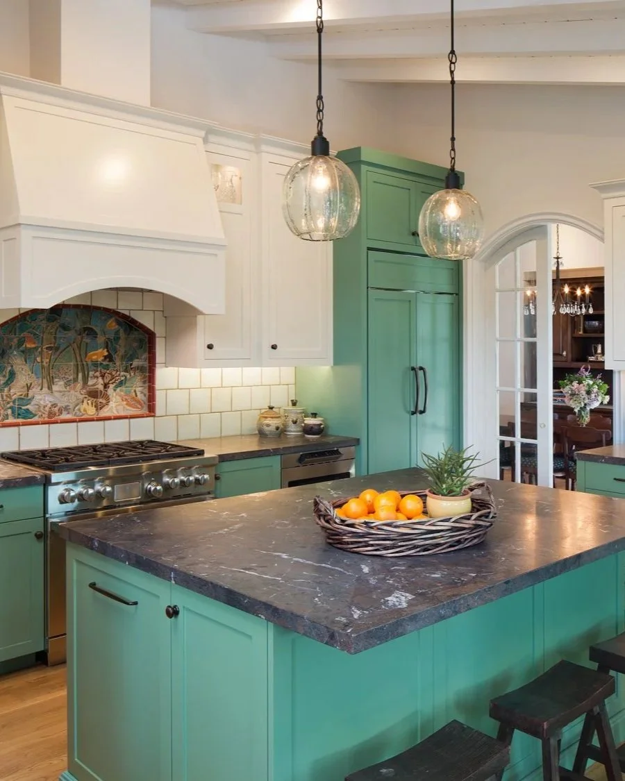 A La Jolla Shores kitchen designed by Tosti Design with green cabinets, a marble countertop, white tile backsplash, and hanging glass pendant lights over the island. There is a basket with oranges and a small potted plant on the island.