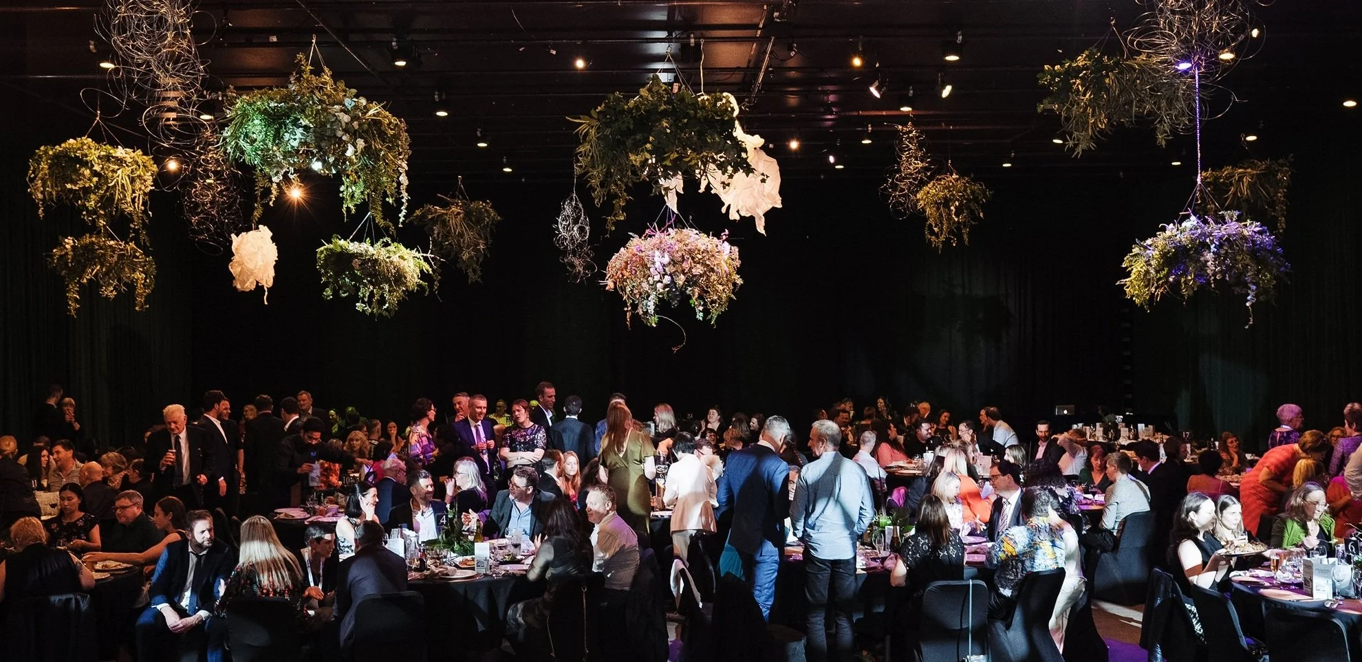Conference attendees networking during a gala dinner beneath decorative floral installations.