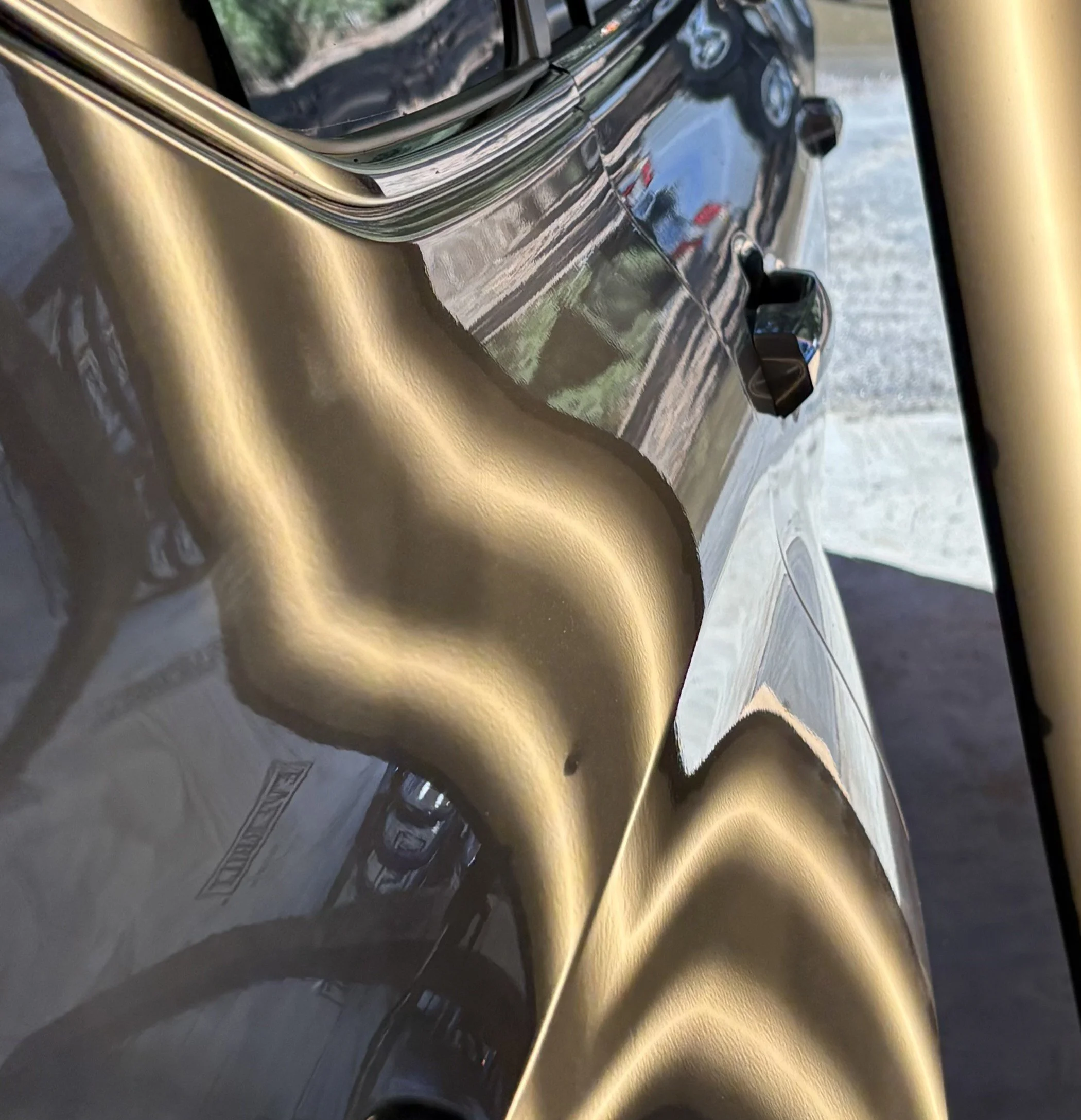 Close-up of a black car with a reflective, gold-colored accent or detail on its body, with a parking lot and other vehicles visible in the reflection.