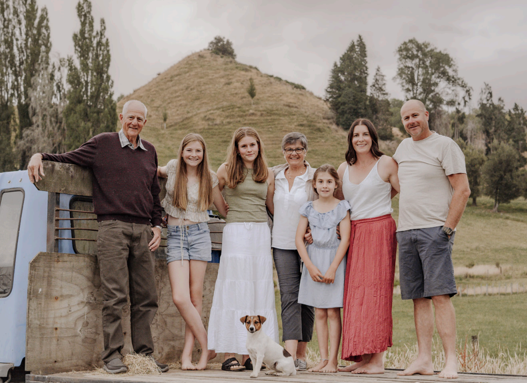 Family of eight, including children, adults, and seniors, standing outdoors on a farm or rural setting near a truck, with trees and a hill in the background.