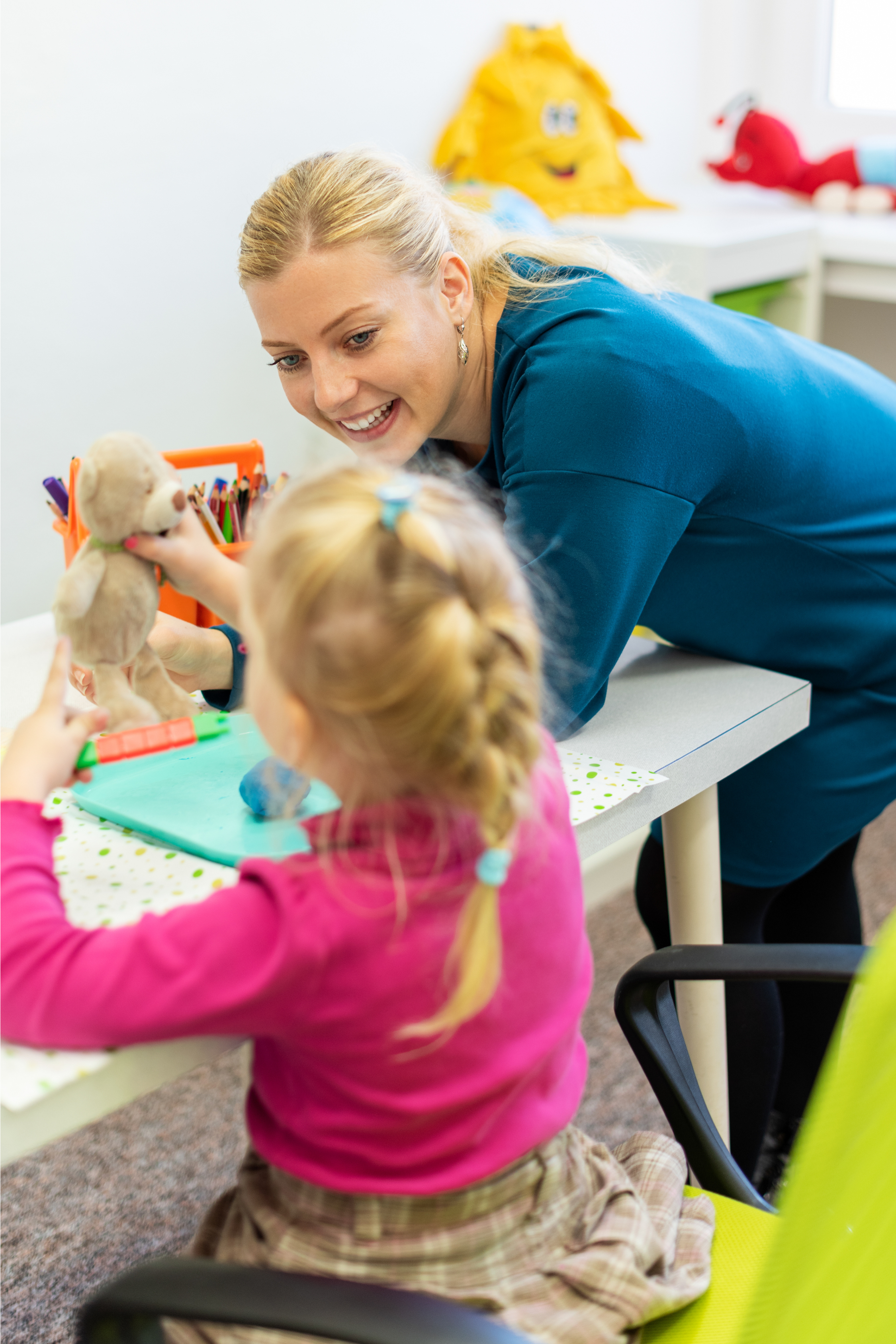A woman in blue scrubs interacting with a young girl at a table, with the girl holding a teddy bear and a toy in her hand.