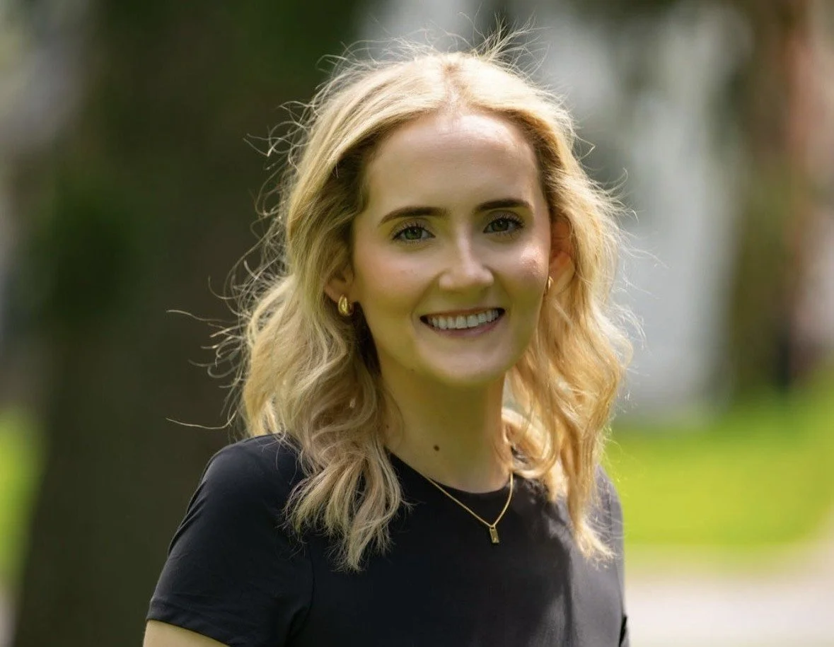 A smiling blonde woman with wavy hair wearing a black top and gold jewelry outdoors.