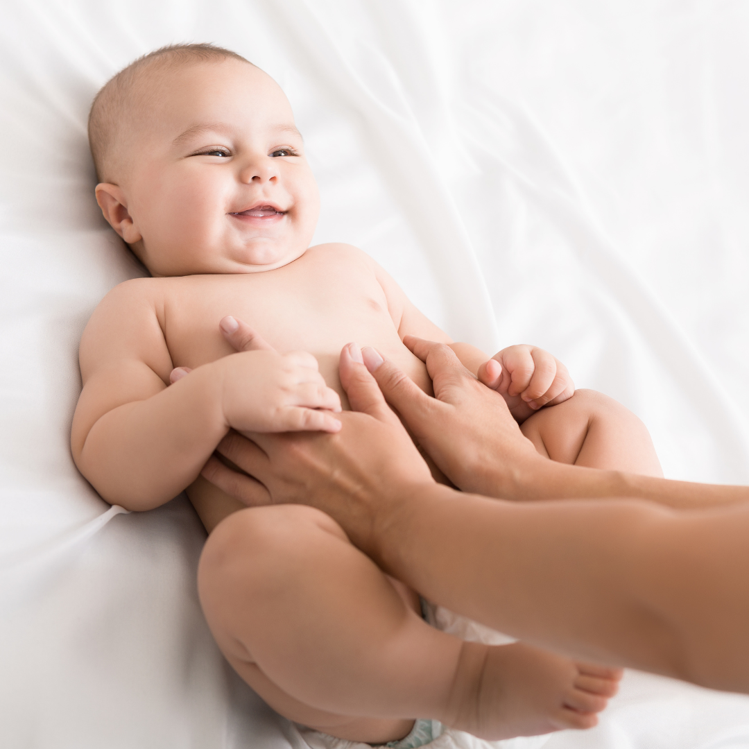 A smiling baby lying on a white sheet while an adult gently touches the baby's stomach.