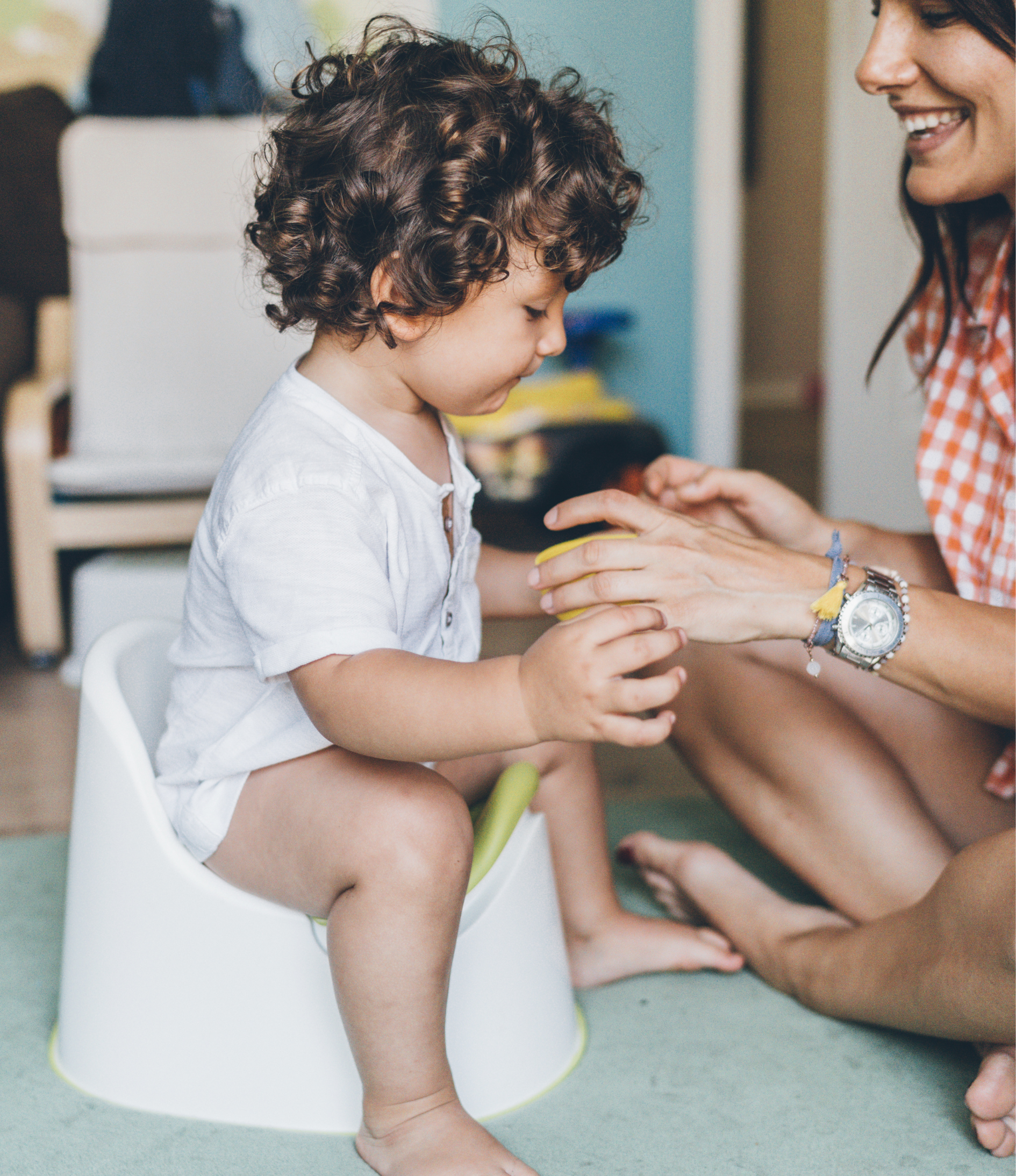 A young child with curly hair sitting on a potty, receiving a treat or small object from a smiling woman, in a colorful, child-friendly room.