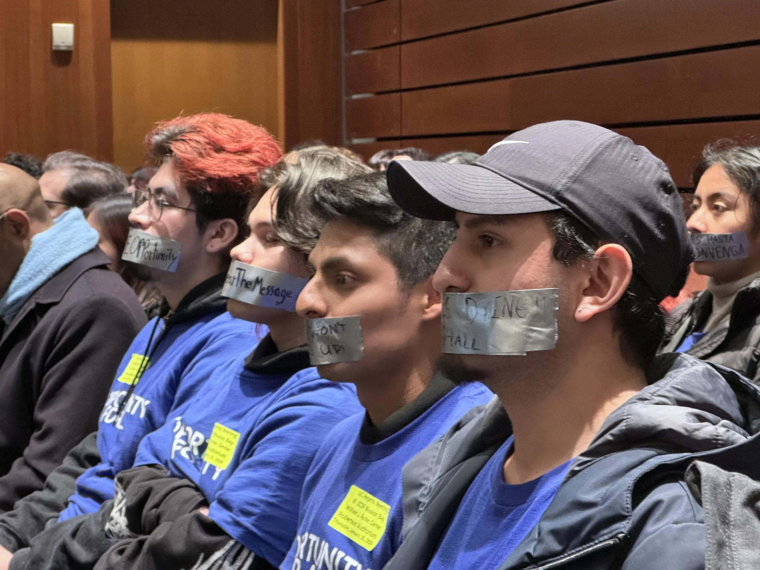 A group of young people sitting side by side at an indoor event, with their mouths covered by pieces of tape with messages written on them.