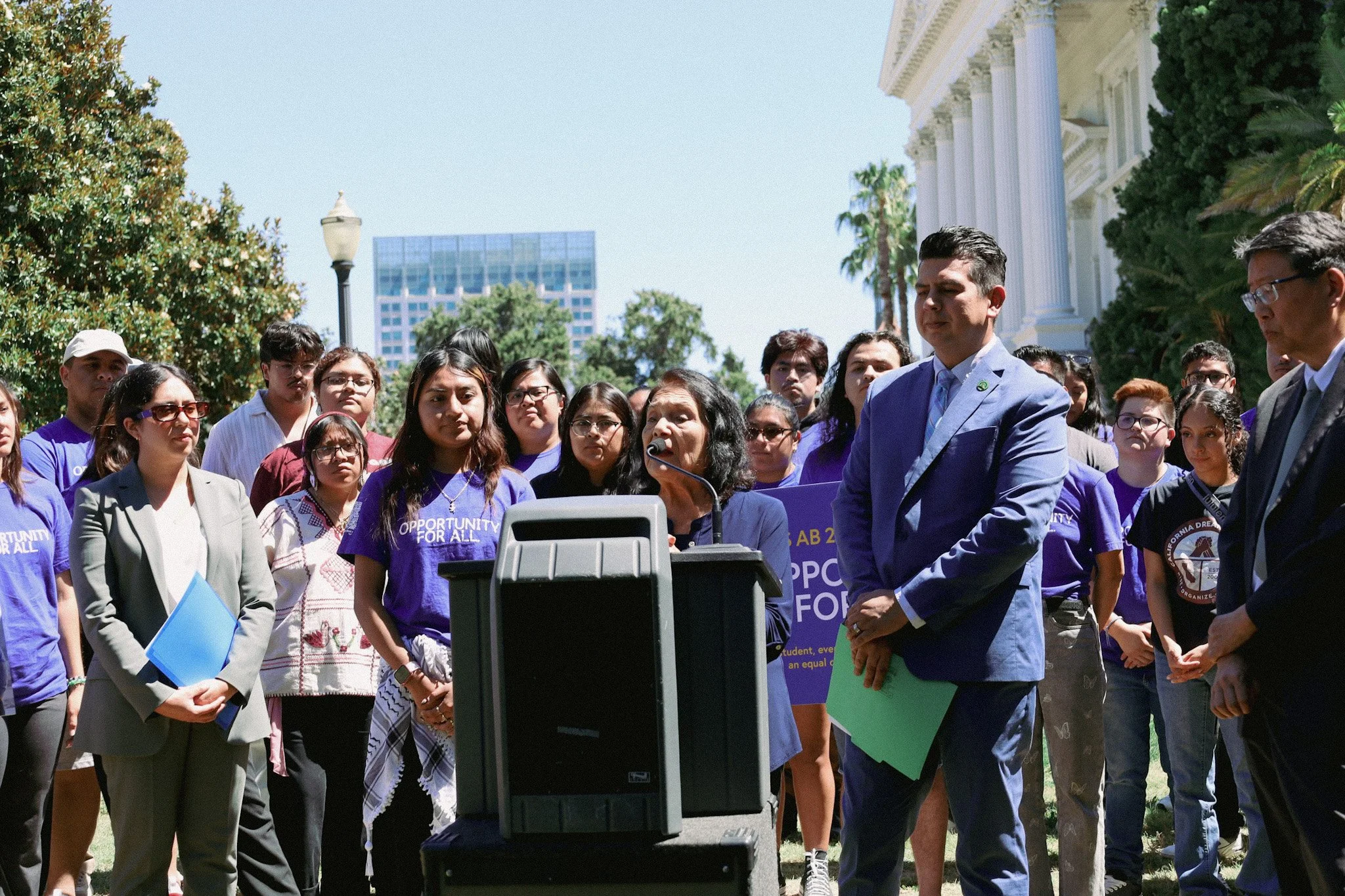 A diverse group of people gathered outdoors for a rally or press conference, with a woman speaking at a podium and others holding signs and listening attentively.