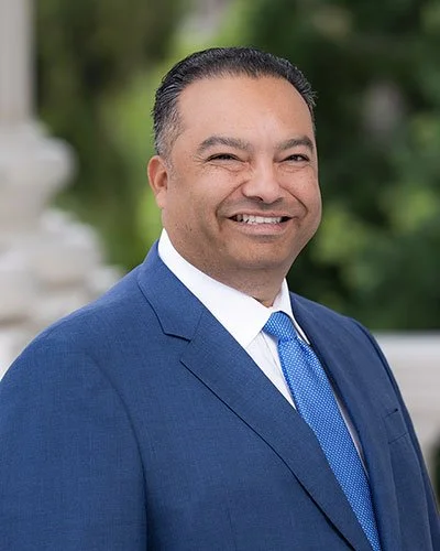 A smiling man in a blue suit with a white shirt and blue tie, outdoor setting with greenery and a white fountain in the background.