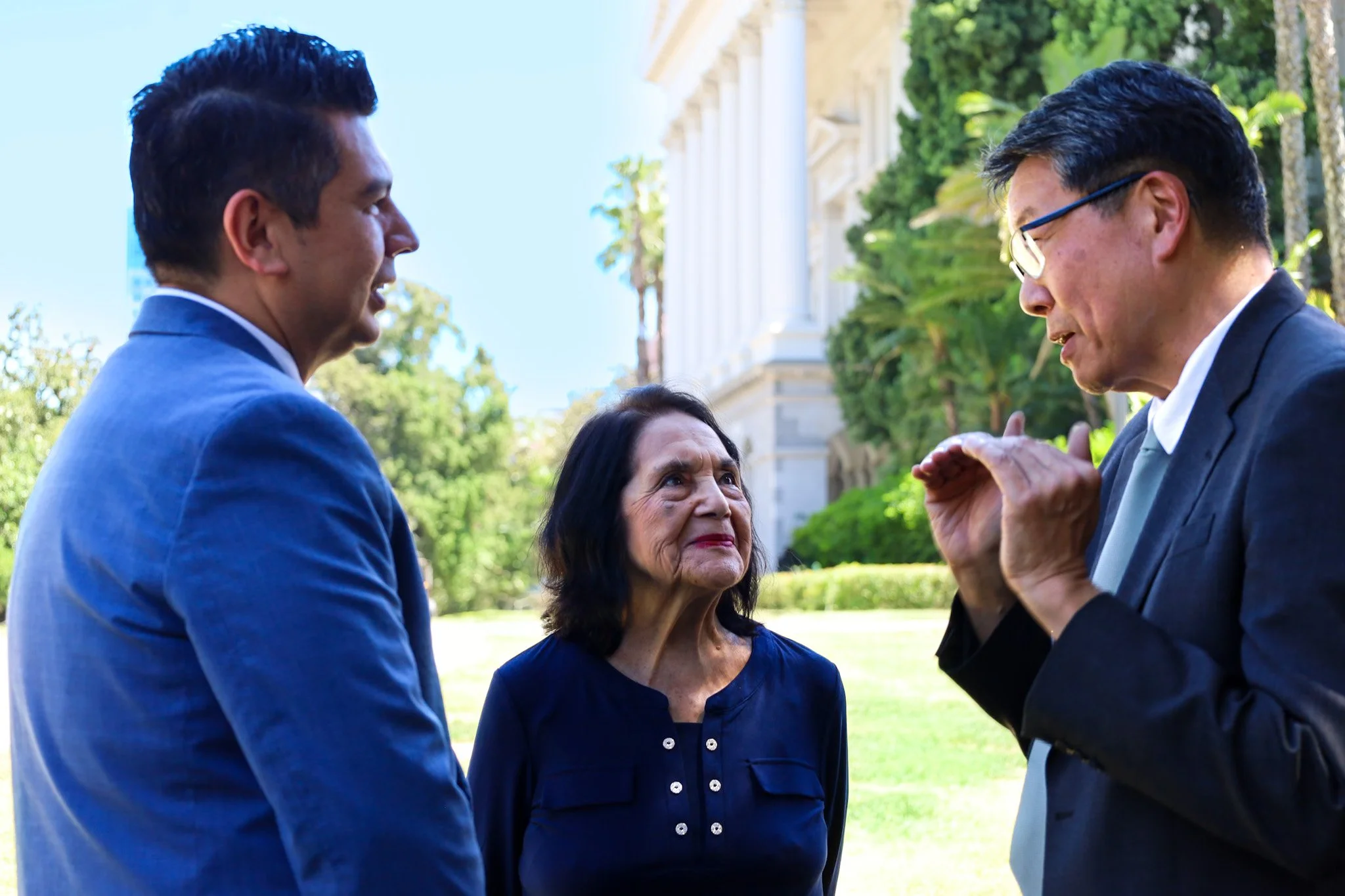 Three people in conversation outdoors, two men and one woman, with a historic building in the background and greenery all around.