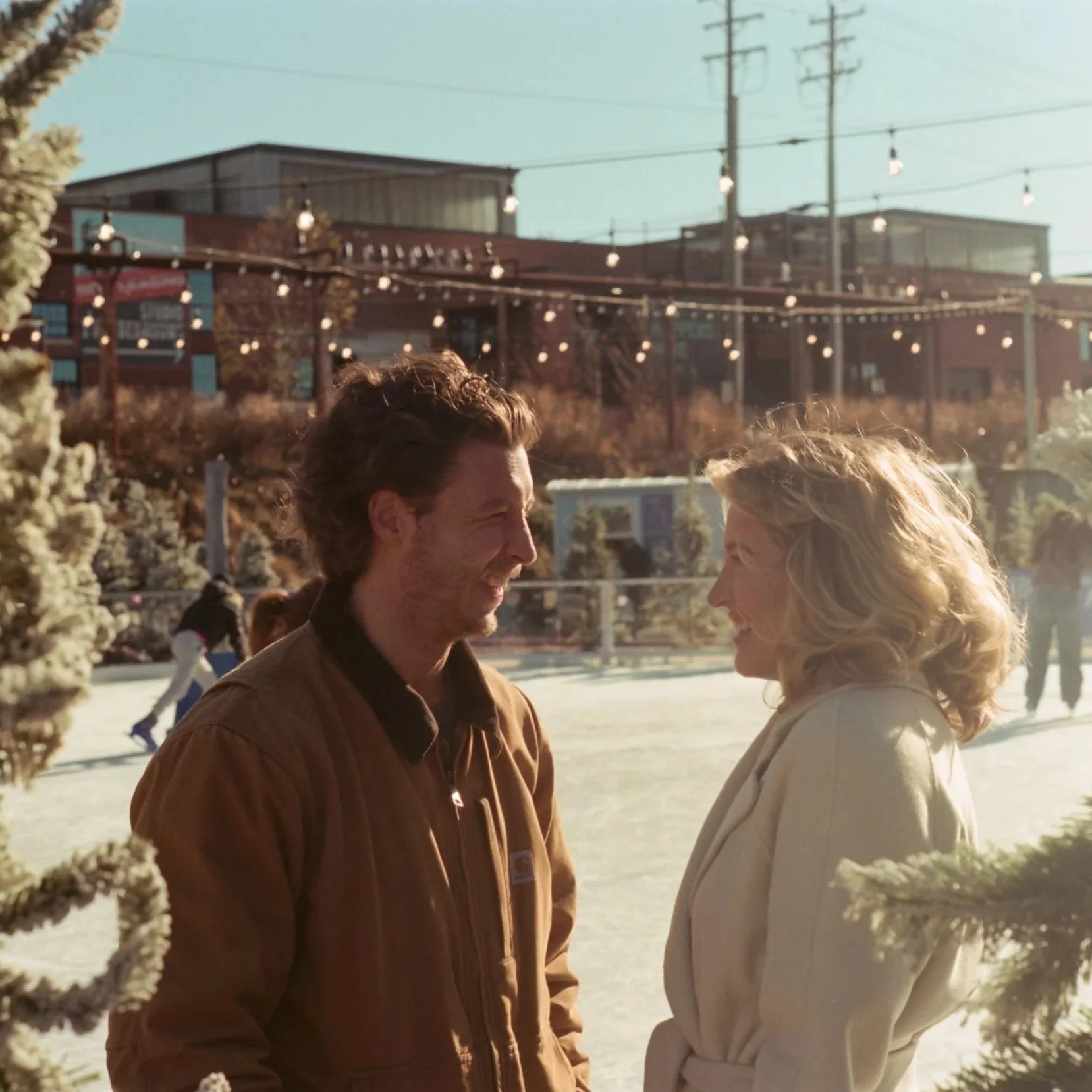 A man and woman smiling at each other on an ice skating rink with holiday lights and decorated trees in the background.
