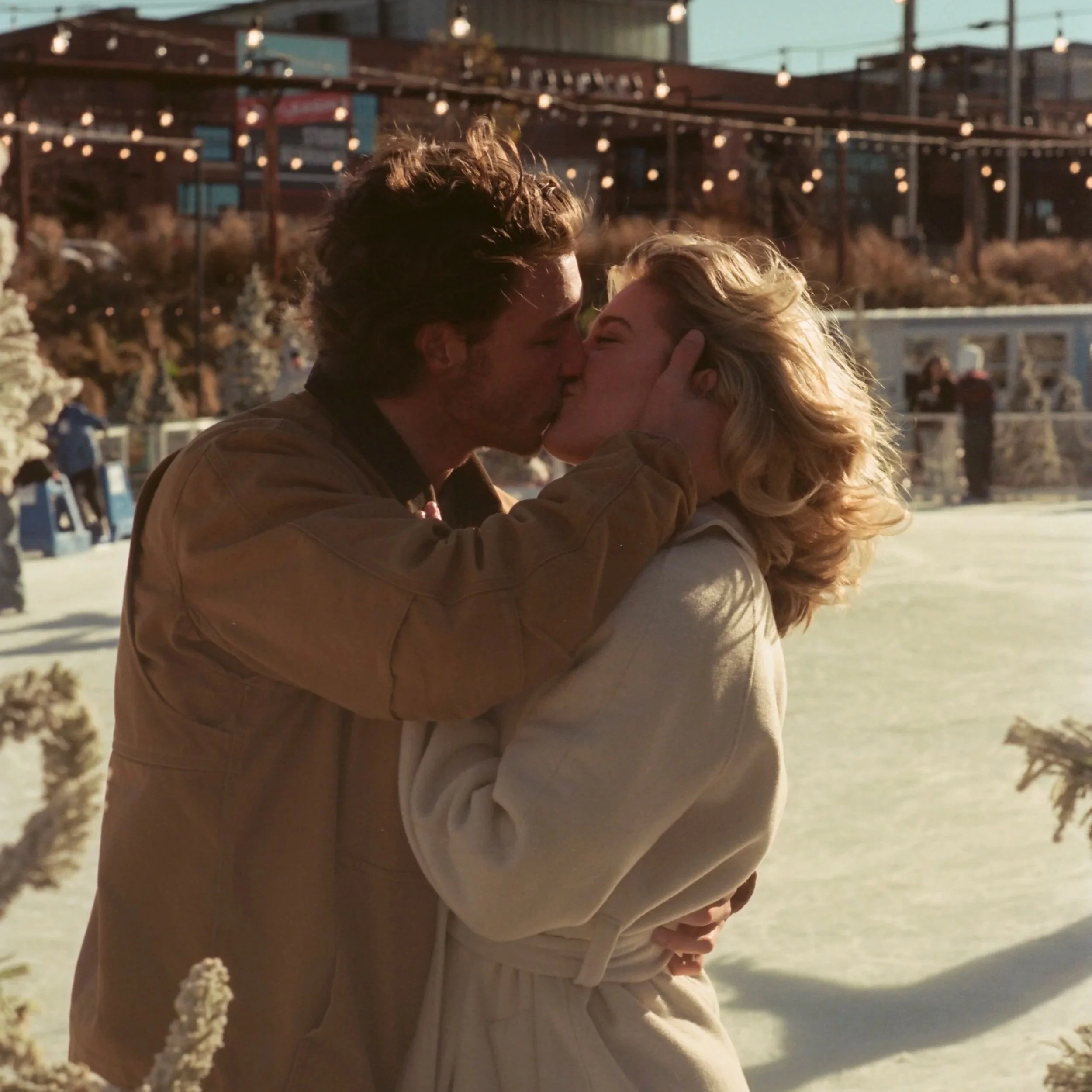 A man and woman sharing a kiss outdoors on a snowy ice rink at sunset, with string lights overhead.