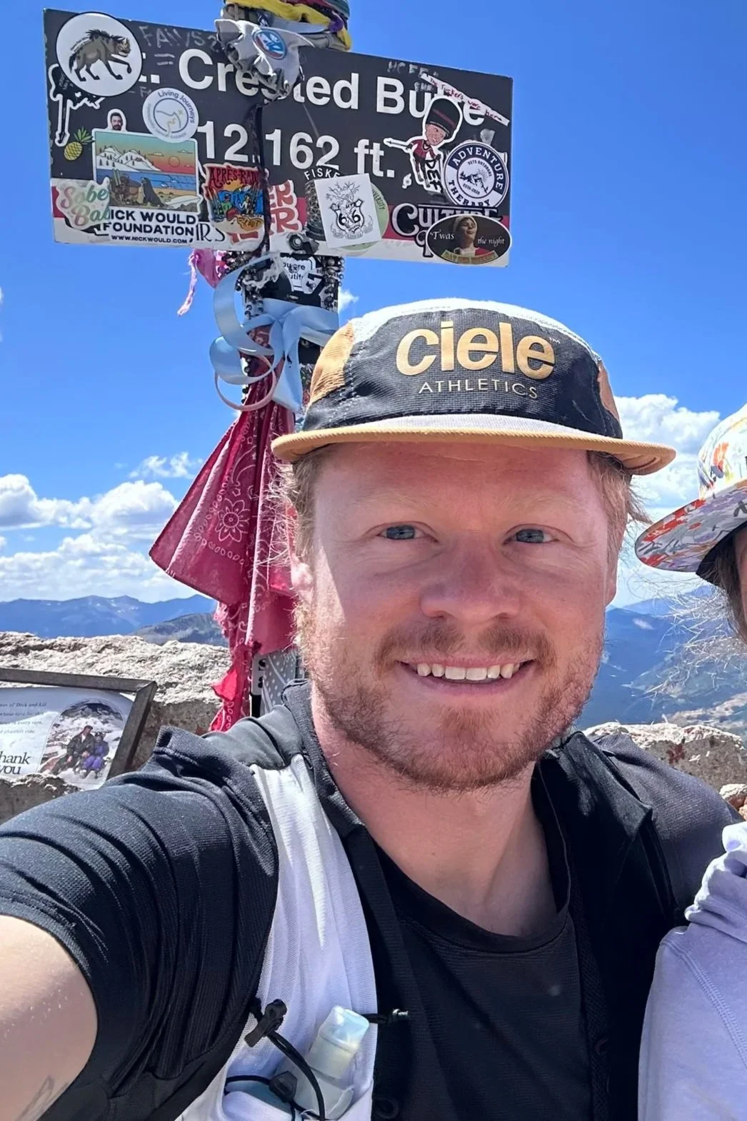 A man with a beard smiling in a mountain setting, wearing a black and white athletic shirt and a tan and black cap with 'Ciele Athletics' written on it. Behind him is a sign indicating an elevation of 12,162 feet, decorated with stickers and ribbons, with mountains and a blue sky with clouds in the background.