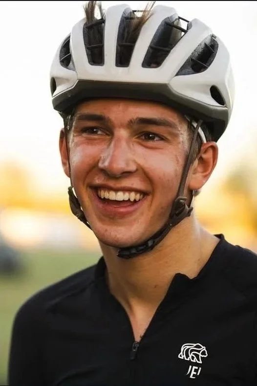 Young man smiling and wearing a white bike helmet and a black zip-up sports jersey with a white logo.