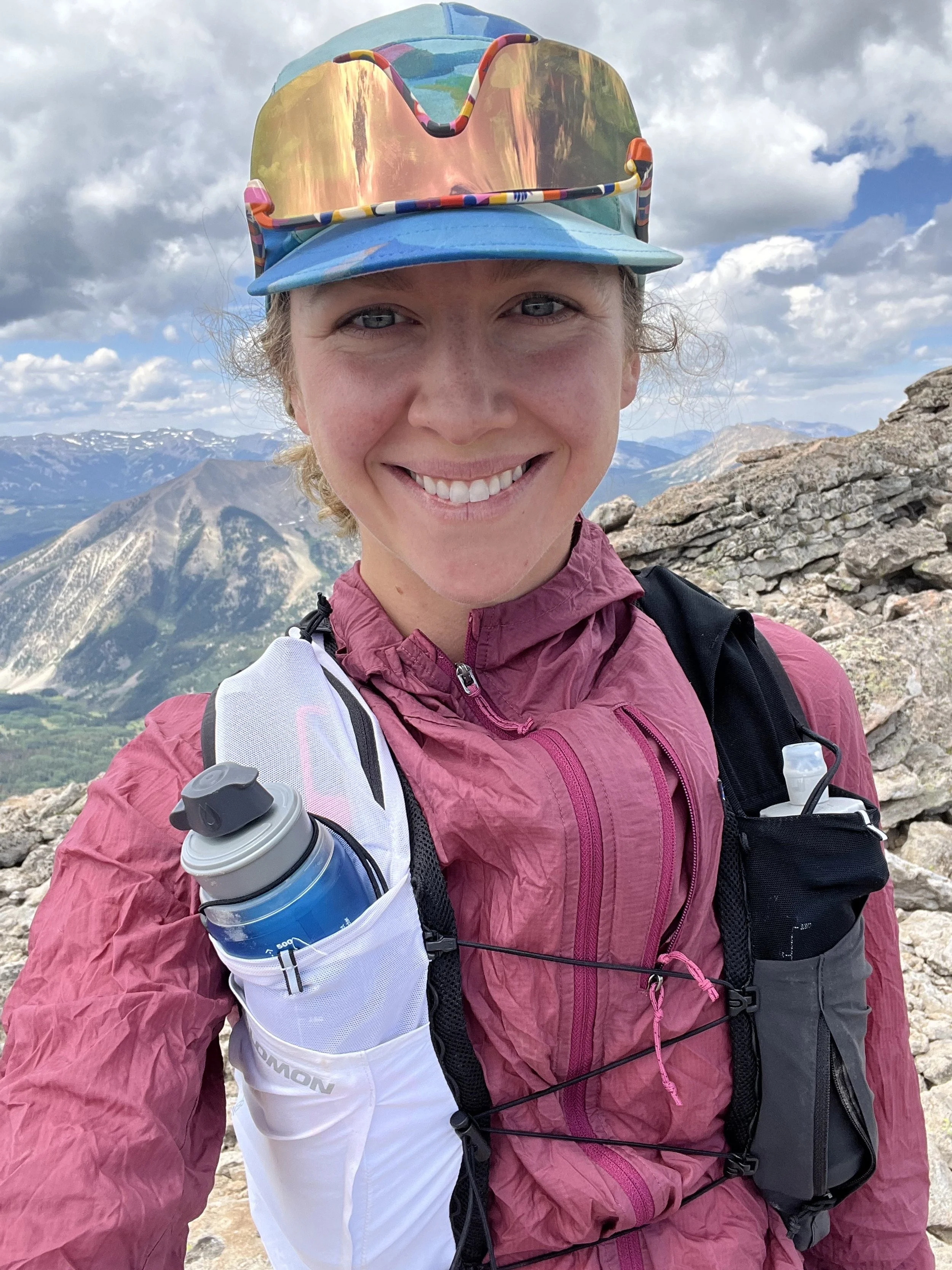 A smiling woman wearing a pink jacket, sunglasses, and a colorful hat, standing on rocky terrain with mountains and a cloudy sky in the background.