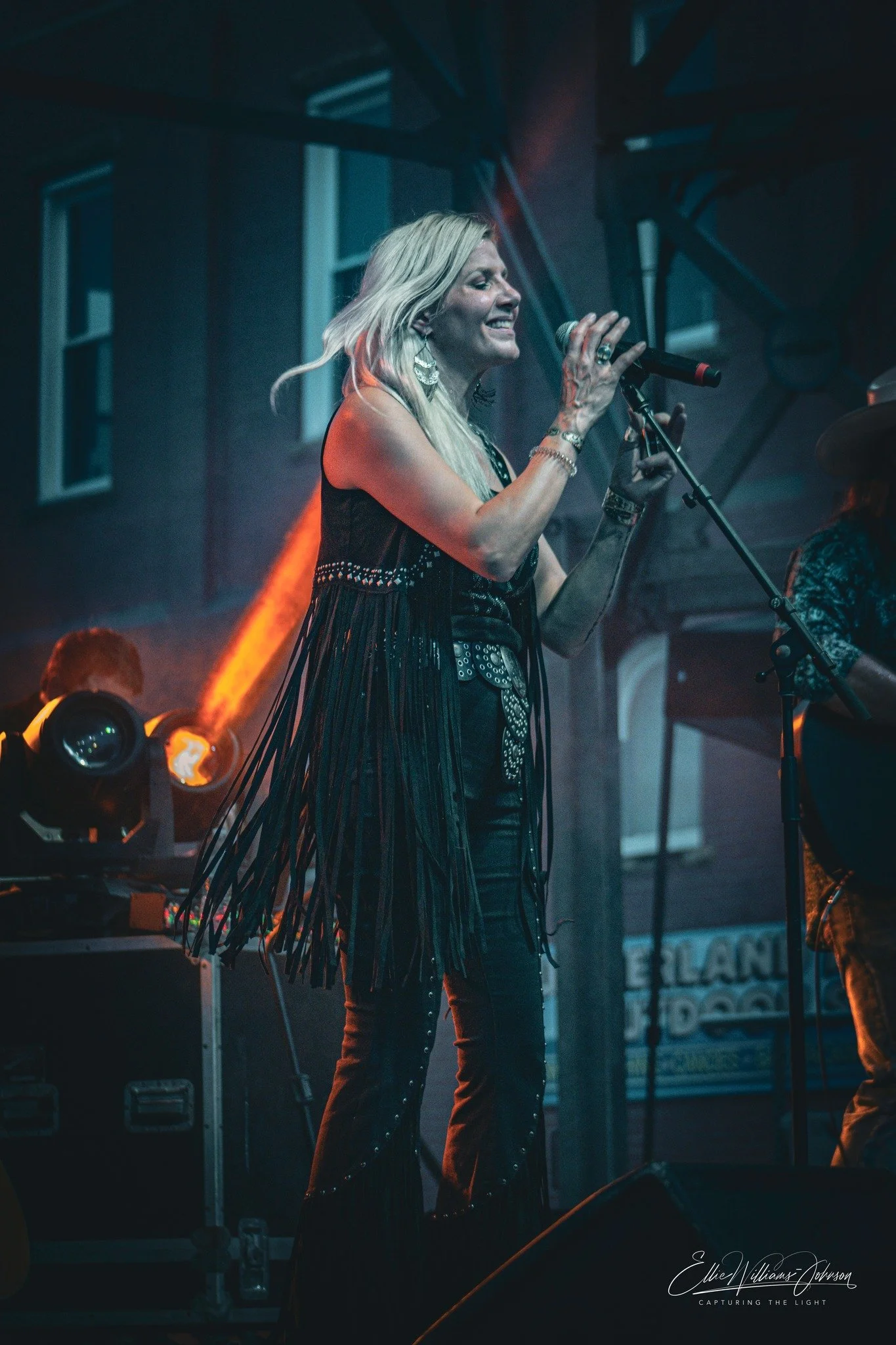 Female singer performing on stage with microphone, wearing fringed black outfit, with stage lights and musicians in the background.