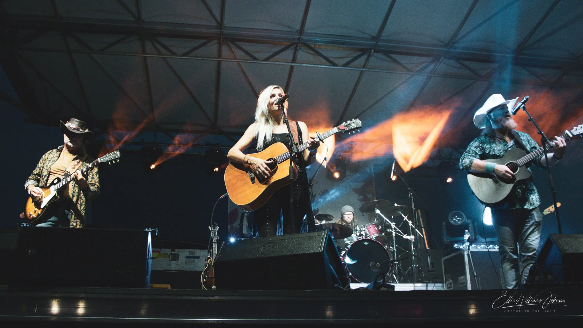 A band performing on stage with three musicians in the foreground playing guitars and singing, and a drummer in the background. Stage lighting includes orange and blue spotlights, and the band members are dressed in country and rock-inspired clothing.