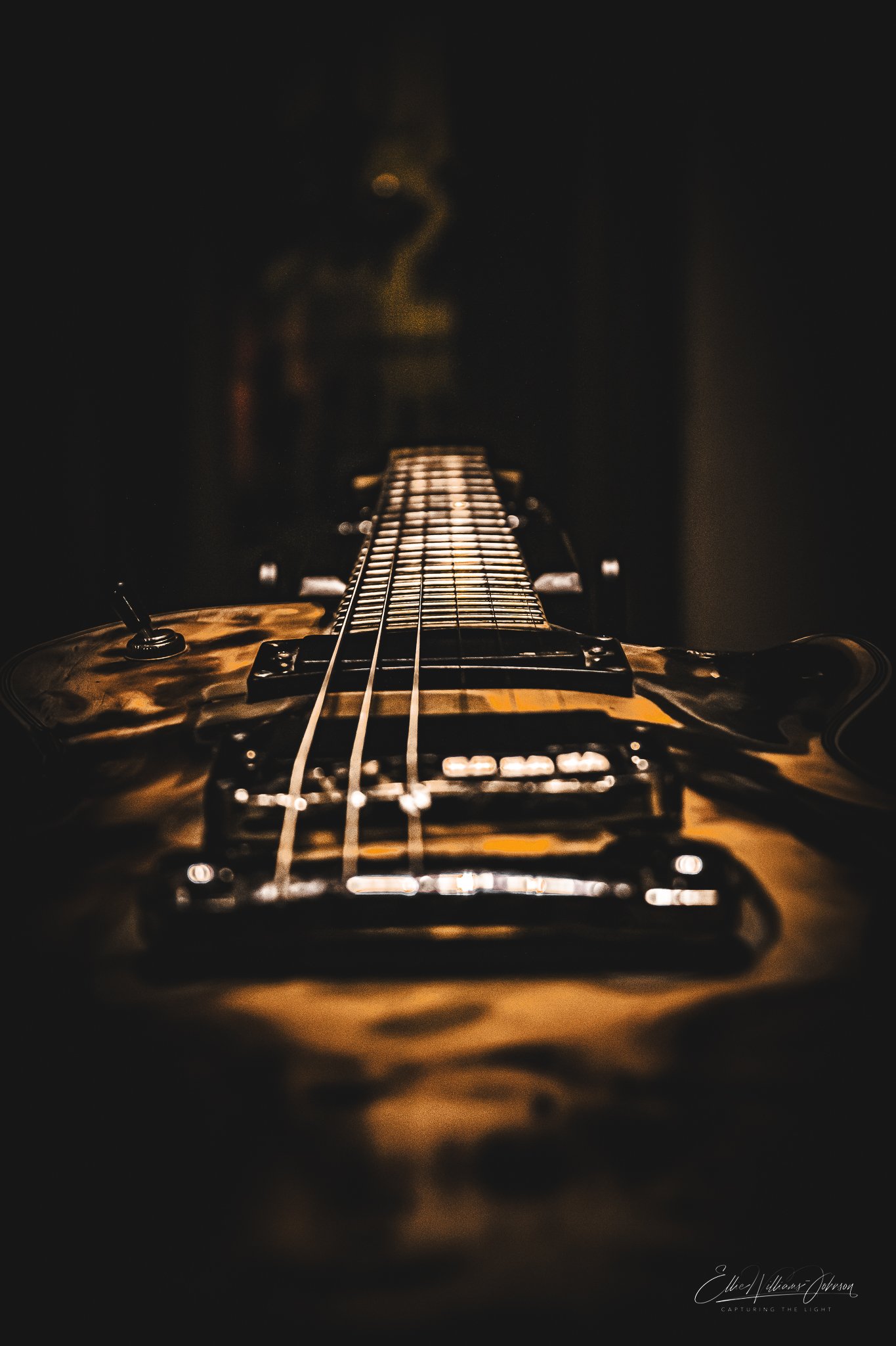 Close-up photo of an electric guitar with a wooden finish, viewed from the body towards the headstock in a dimly lit setting.
