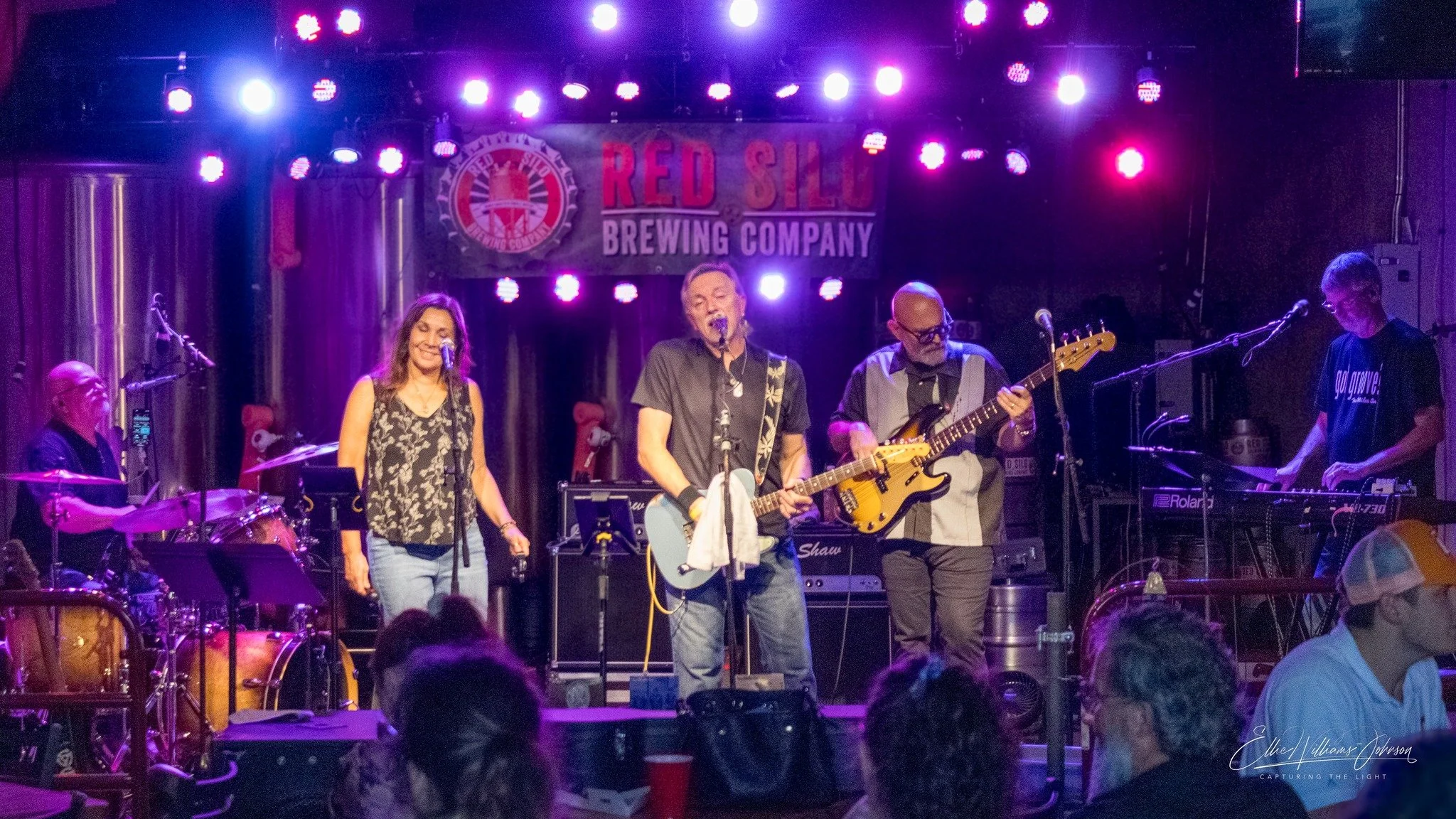 Band performing live on stage at Red Shoot Brewing Company with four musicians, including a man singing and playing guitar, a woman singing, a man playing bass, and a man on keyboard, with colorful stage lights and audience in foreground.