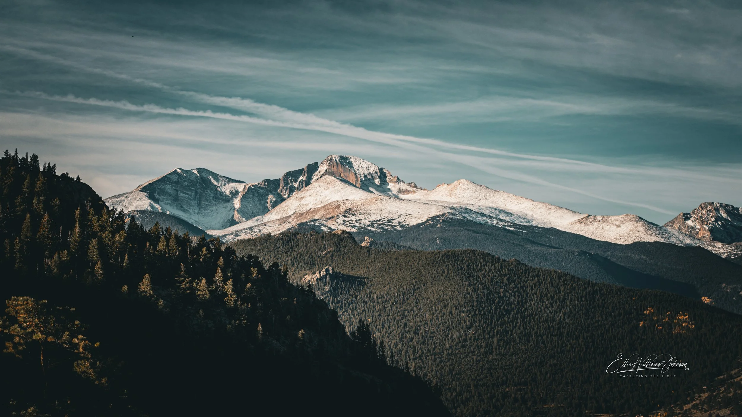 Snow-capped mountain peaks over a lush green forest with a blue sky and wispy clouds.