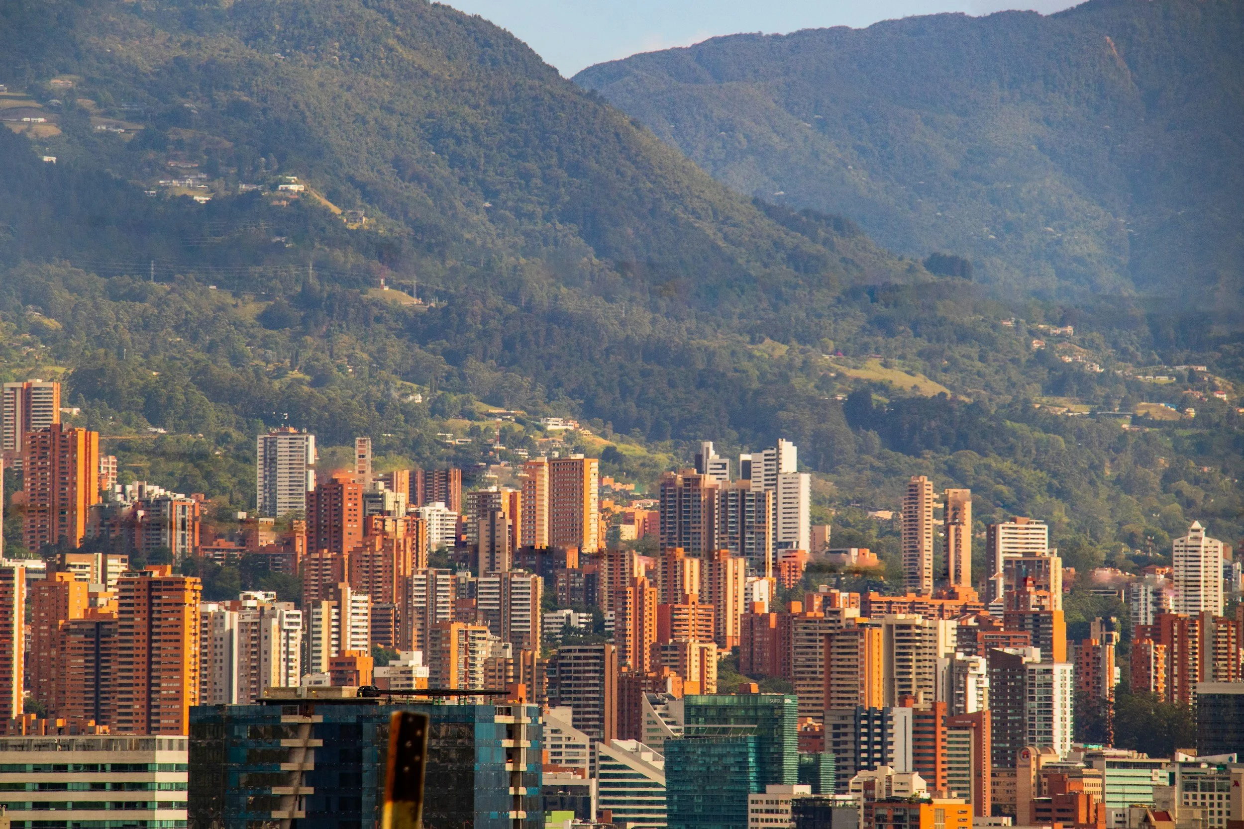 City skyline with tall apartment buildings and skyscrapers in the foreground, with lush green hills and mountains in the background.
