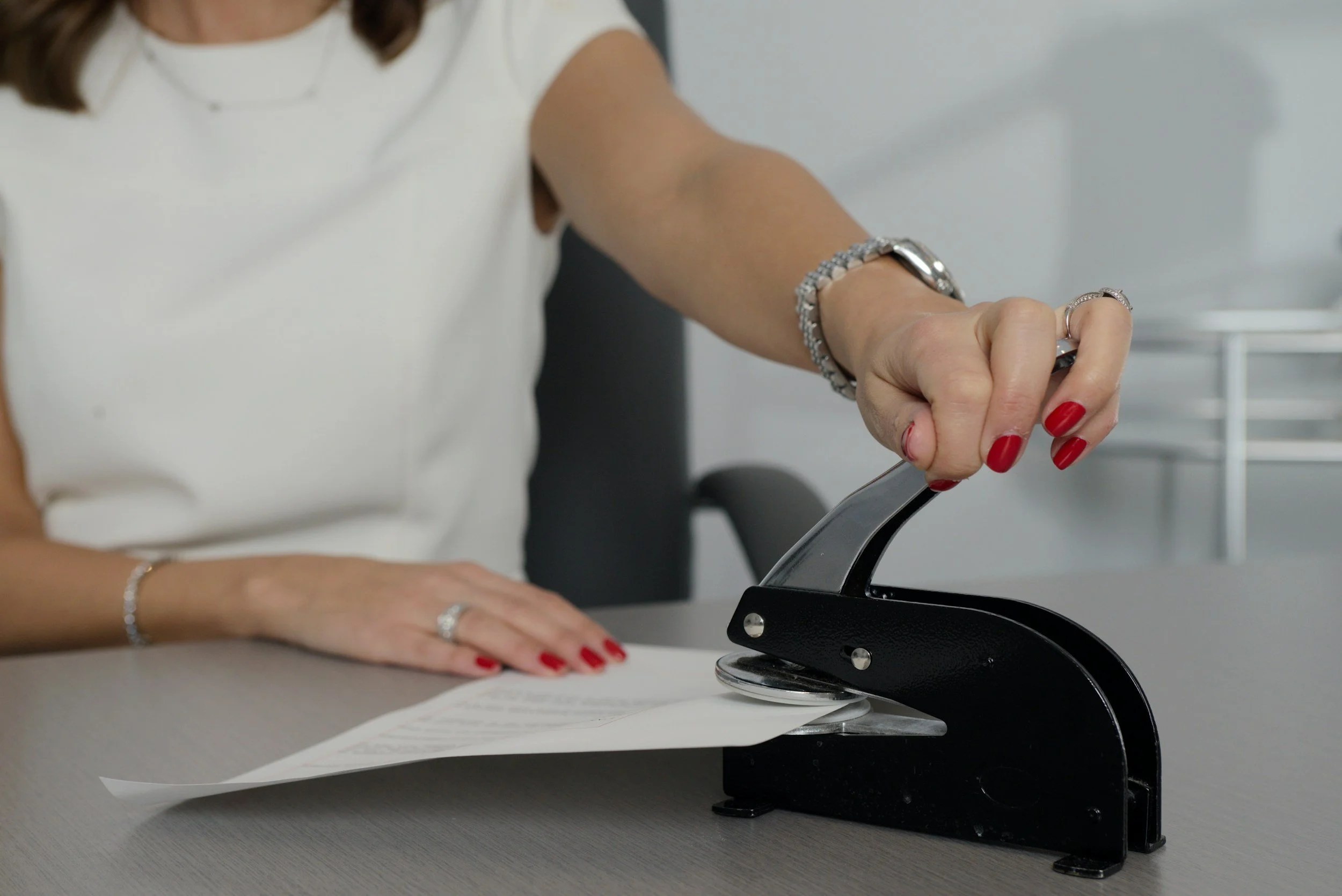 A woman with red painted nails using a hole puncher on a paper sheet in an office setting.