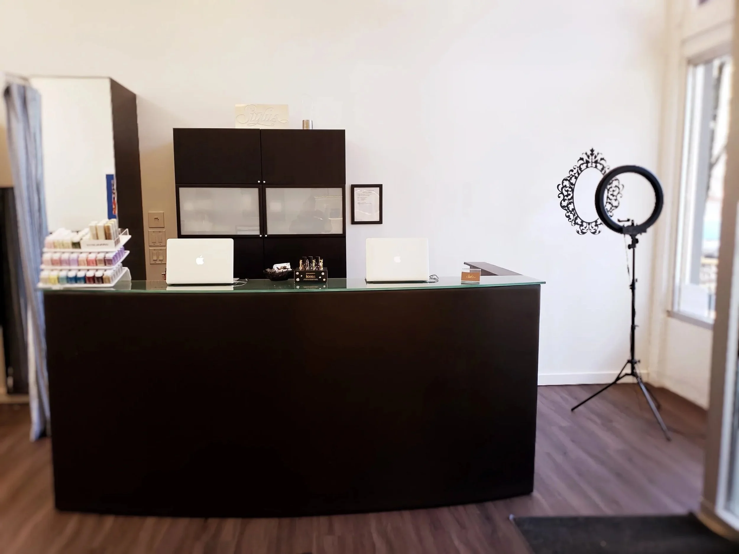 Interior of a nail salon reception area with a black reception desk, two white laptops, and nail polish displays. A ring light is set up to the right of the desk, and a window with blinds is on the far right.