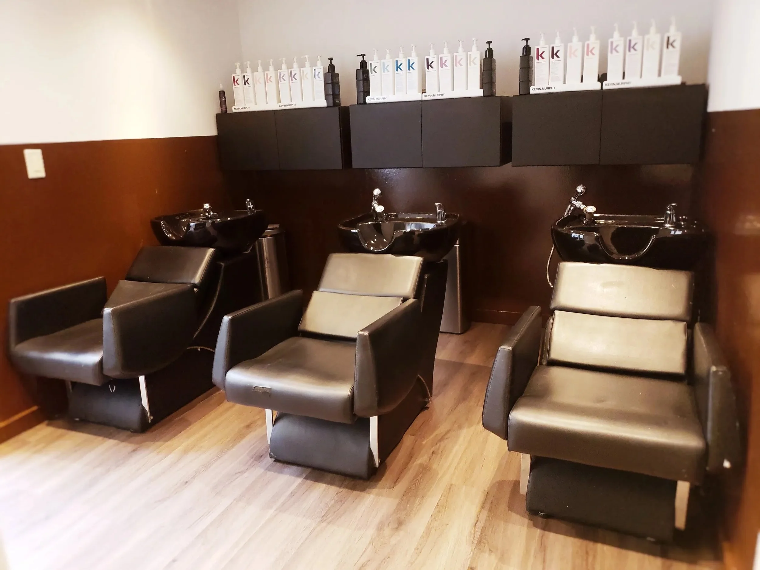 Three black salon chairs with washbasins and black sinks, positioned in a hair salon with brown wood floors and walls, and black cabinets above with hair care products.