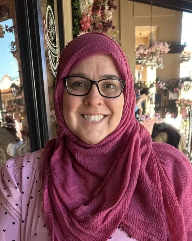 A woman wearing glasses and a pink hijab smiles for a selfie inside a cafe with floral decorations and outdoor seating visible through the window.