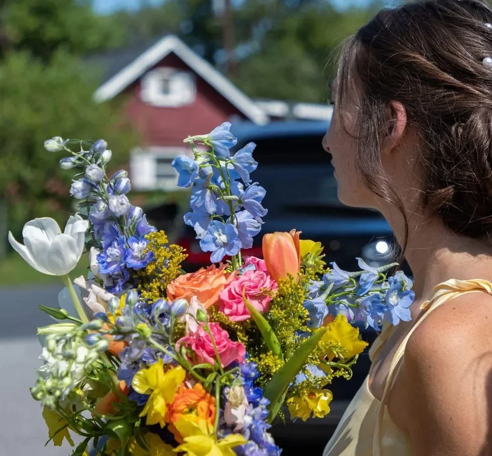 A woman holding a colorful bouquet of flowers, with a house and greenery in the background.