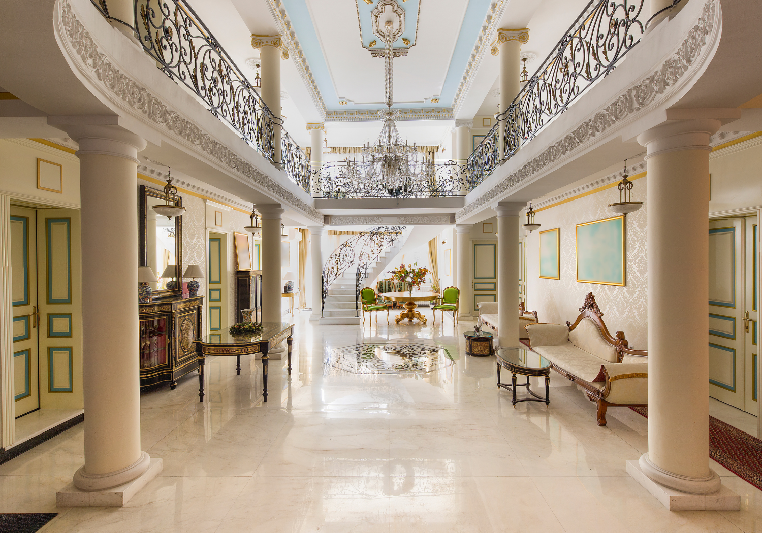 Elegant, luxurious hotel lobby with white marble floors, classical furnishings, and ornate iron railing on the upper level. There are columns, a grand staircase, and a chandelier hanging from the ceiling. The space is decorated with green chairs, paintings, and decorative flowers.