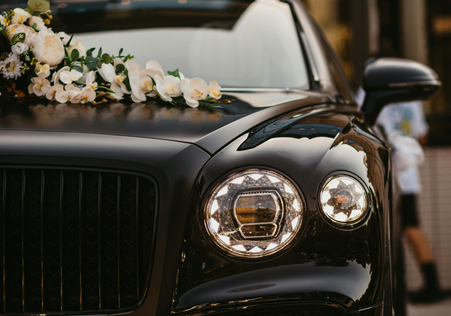 Close-up of a black luxury car with a floral decoration on the hood, featuring white flowers and greenery, during daytime.