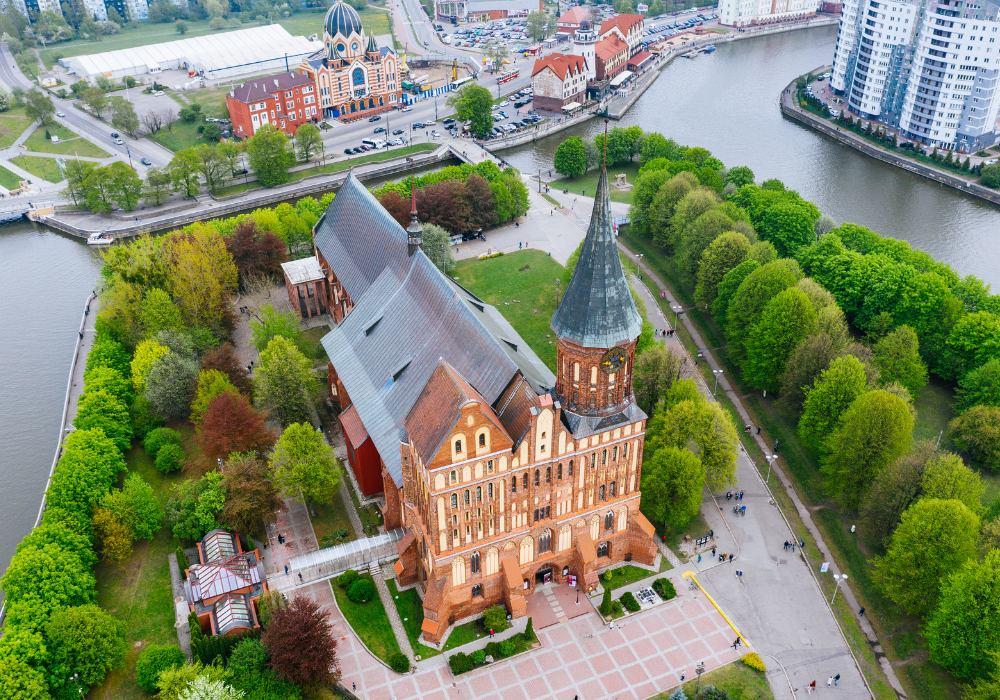 Aerial view of a historic brick church with a tall steeple, surrounded by lush green trees and a park, with a river and modern buildings in the background.