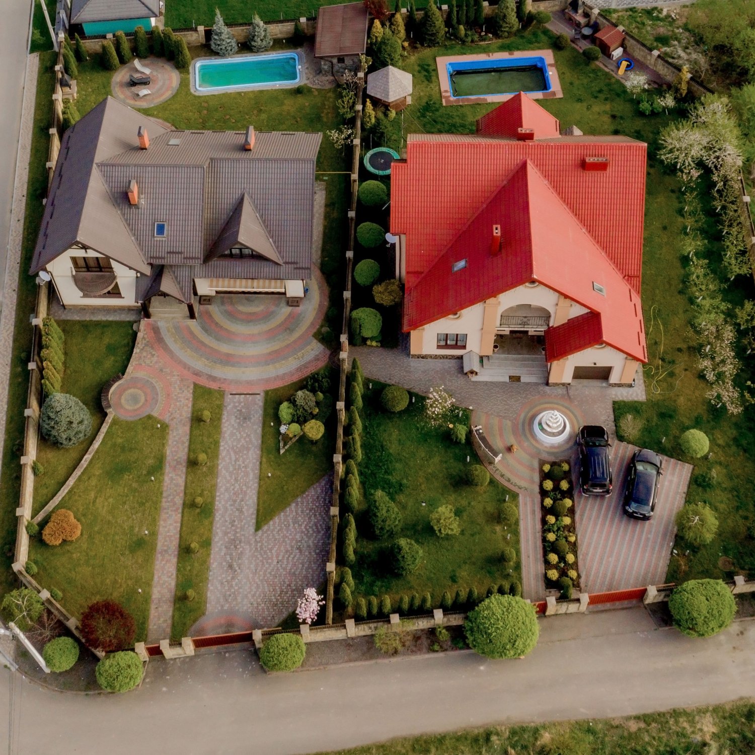 Aerial view of two houses with landscaped gardens, driveways, and swimming pools, surrounded by a fenced yard.
