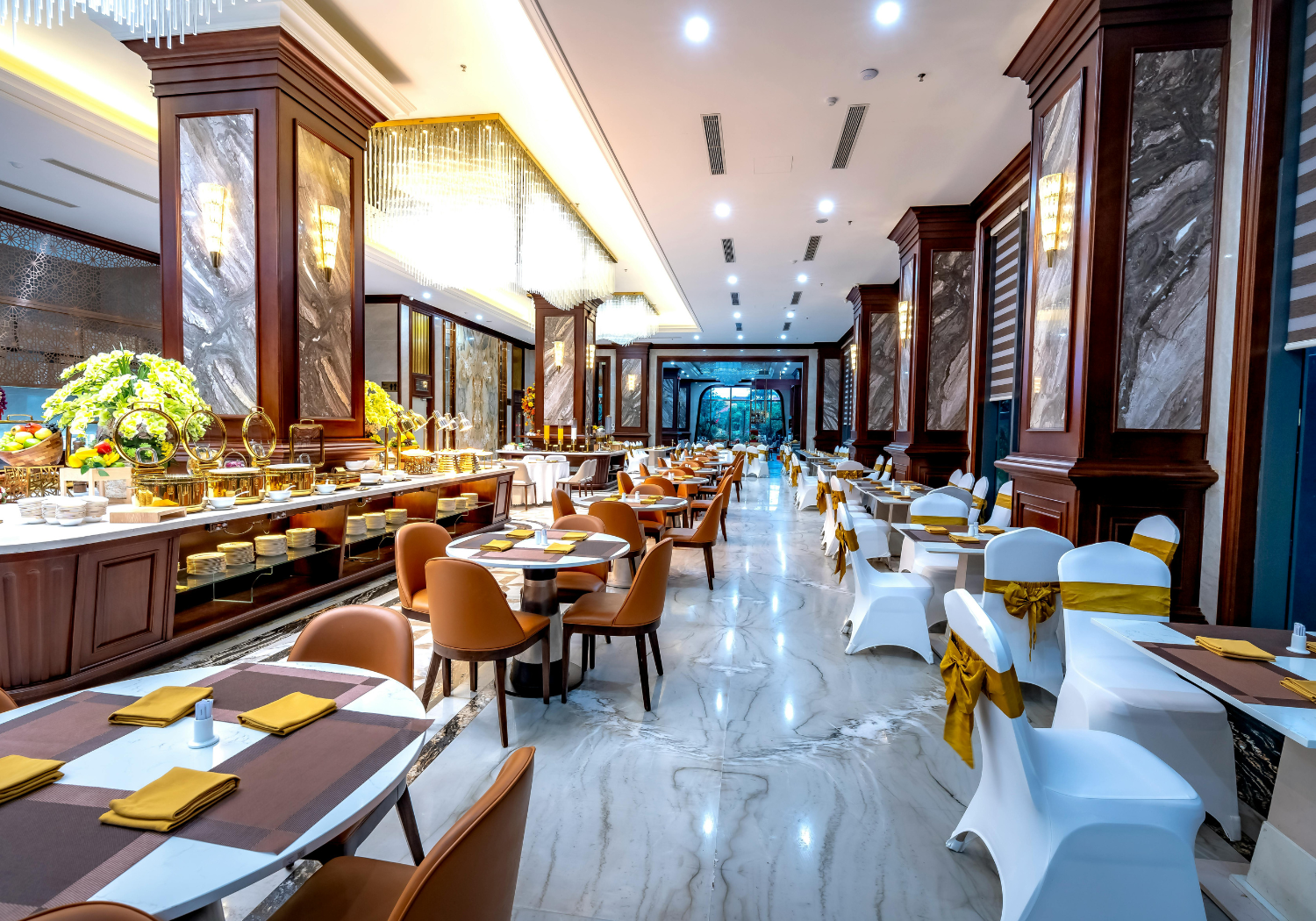 Elegant restaurant interior with round tables set with yellow napkins, chairs covered with white chairs and golden bows, and a buffet table with food and floral decorations. The room features marble flooring, wooden wall panels, and modern lighting fixtures.