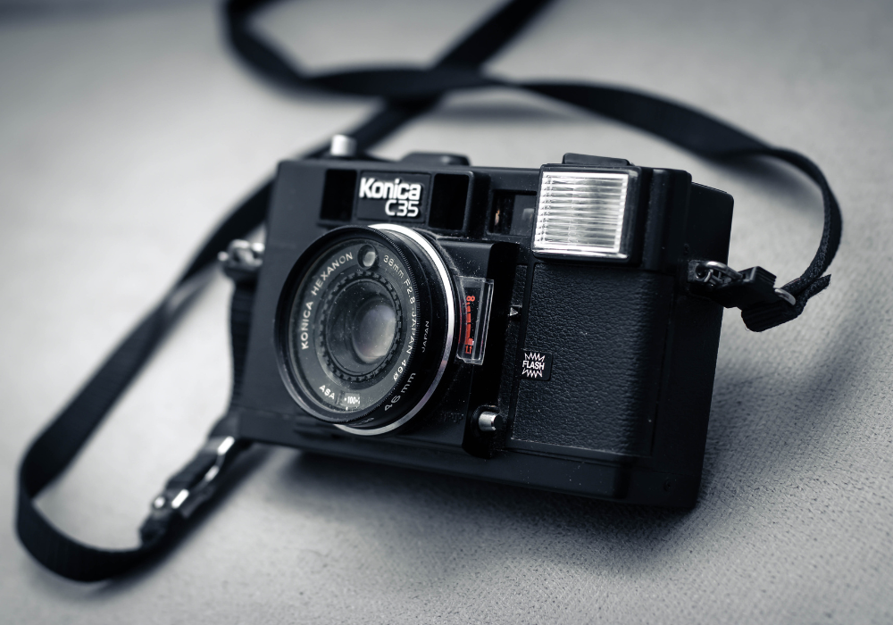 A vintage Konica C35 film camera with a built-in flash, placed on a gray surface with a strap attached.