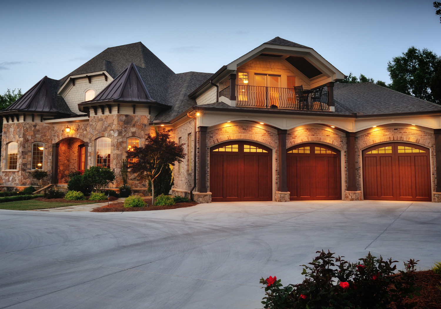 A large, upscale house at dusk with stone and brick exterior, three brown garage doors, and front yard with bushes and a small tree, illuminated by warm exterior lighting.