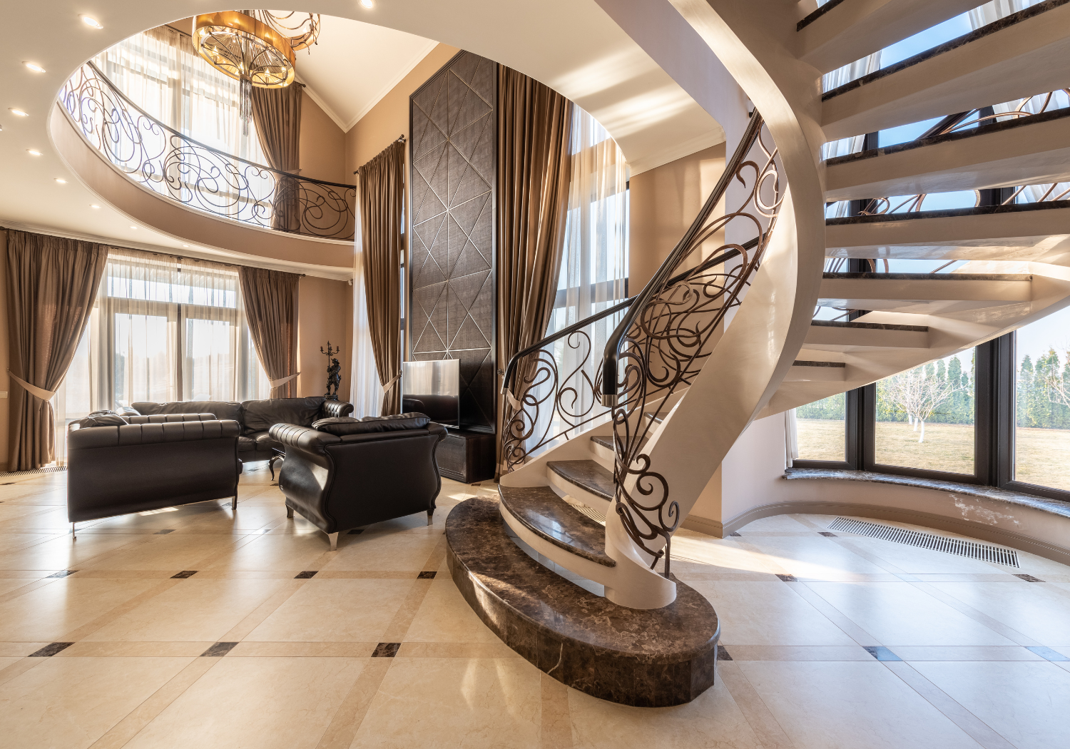 Interior view of a spacious living room with a curved staircase, large windows with beige curtains, and black leather couches.