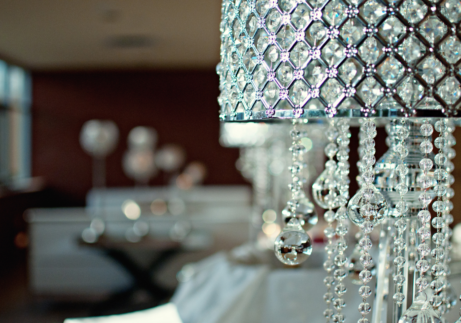 Close-up of a chandelier with crystals and beads hanging from a decorative metal frame, with a blurred background of a dining room with tables and chairs.