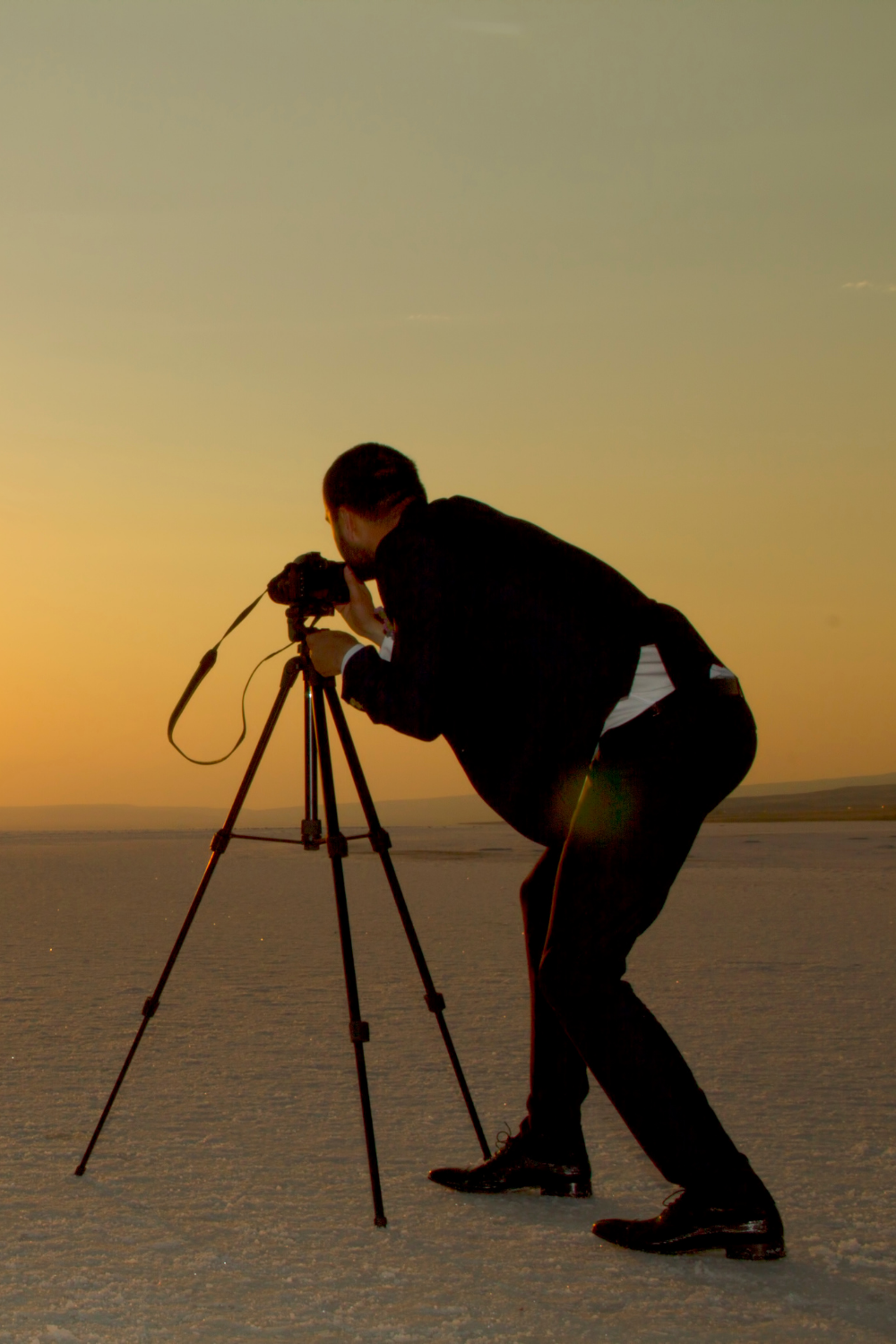 A person in a black suit taking a photograph using a camera on a tripod during sunset over a salt flat.