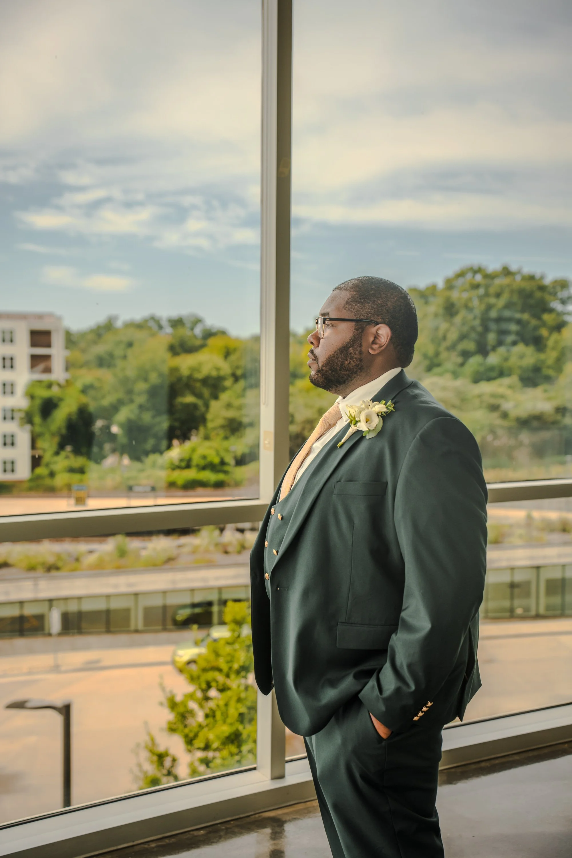 A man dressed in a formal suit with a boutonnière, looking thoughtfully out of a large window at a cityscape with trees and buildings on a partly cloudy day.