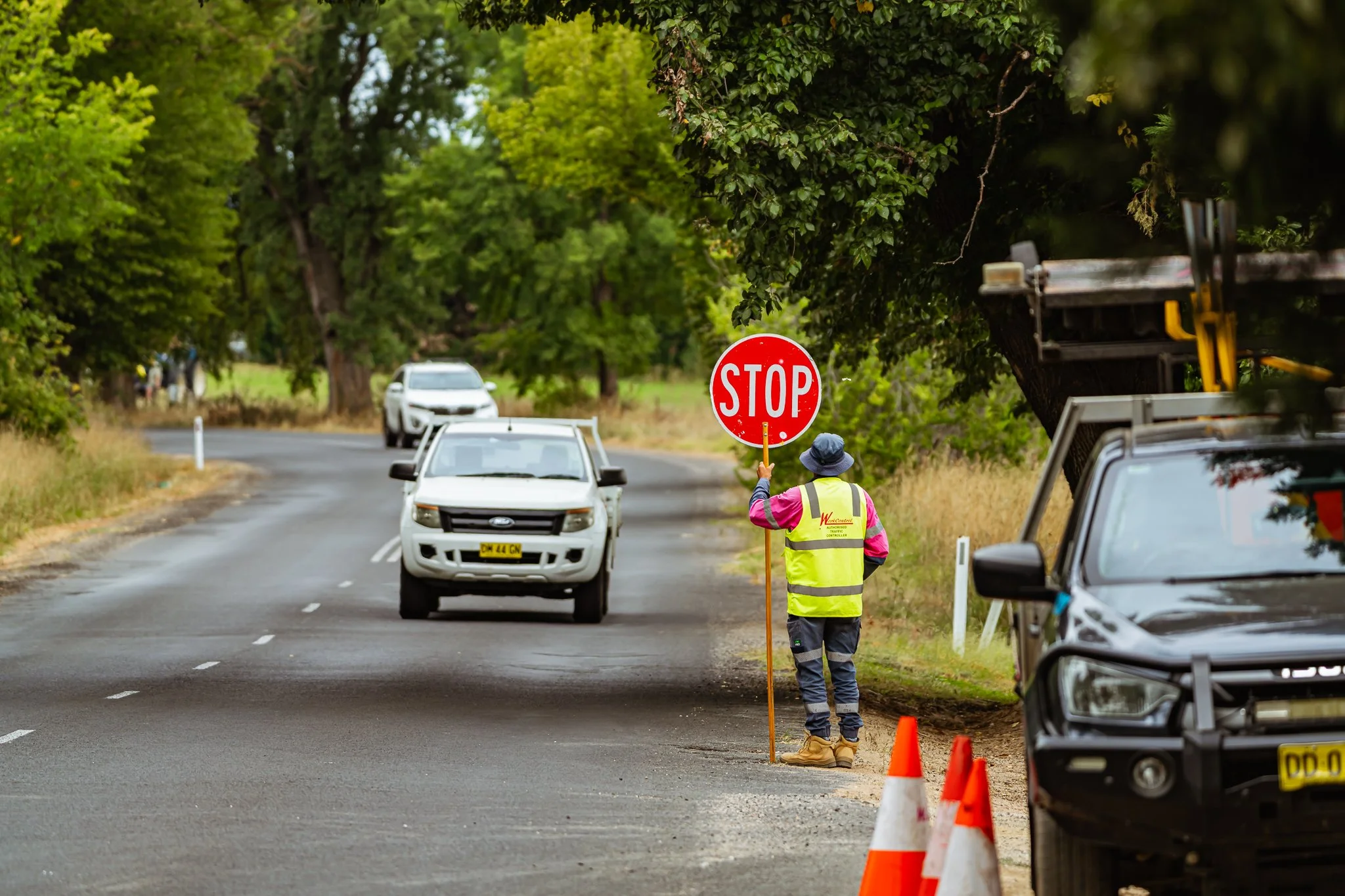 A person wearing a neon safety vest and blue hat holding a stop sign on the side of a rural road, with cars approaching and orange traffic cones nearby.