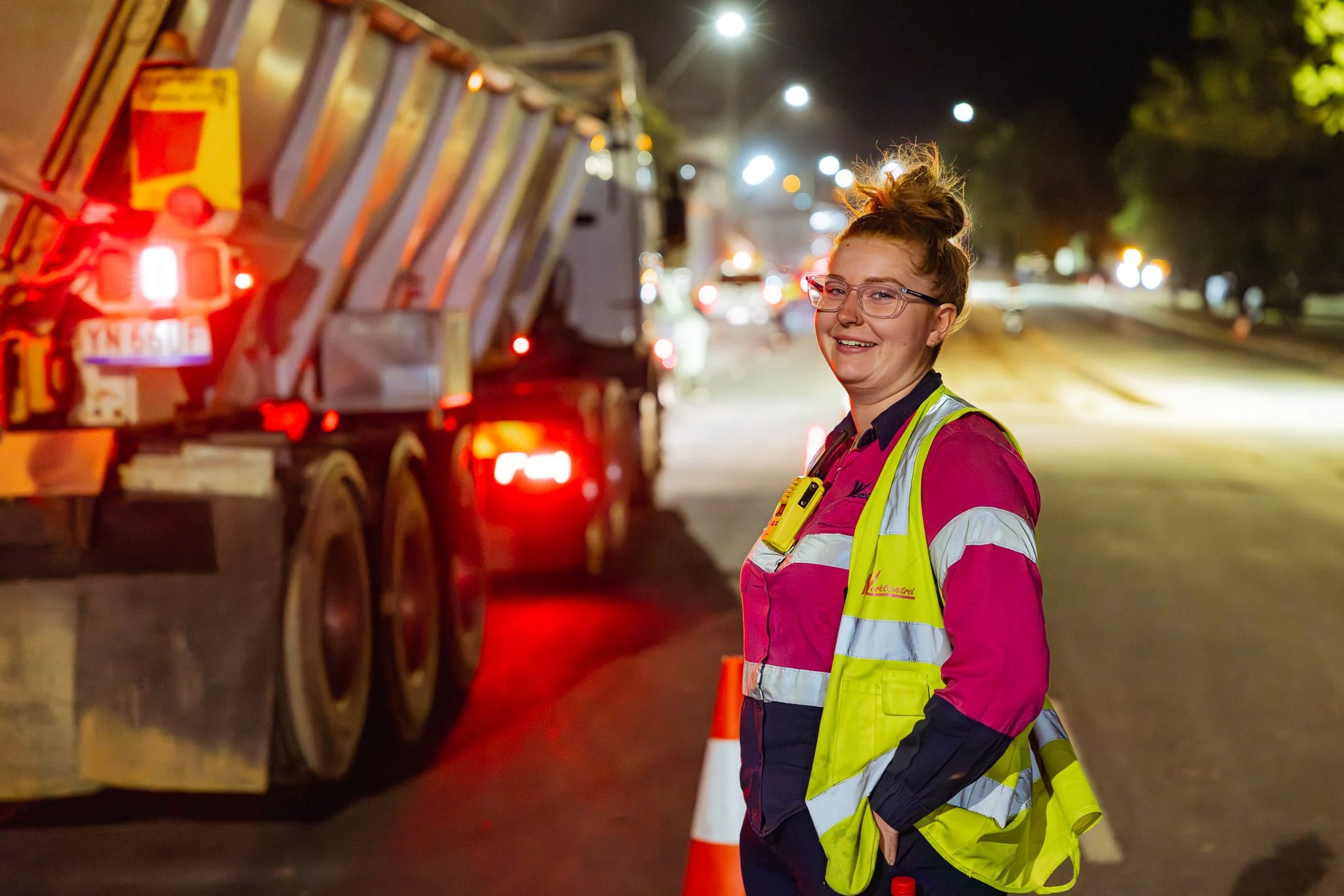 A smiling female construction worker in a pink shirt and yellow safety vest with reflective stripes standing on a nighttime road near a large truck, with traffic cones and bright streetlights in the background.