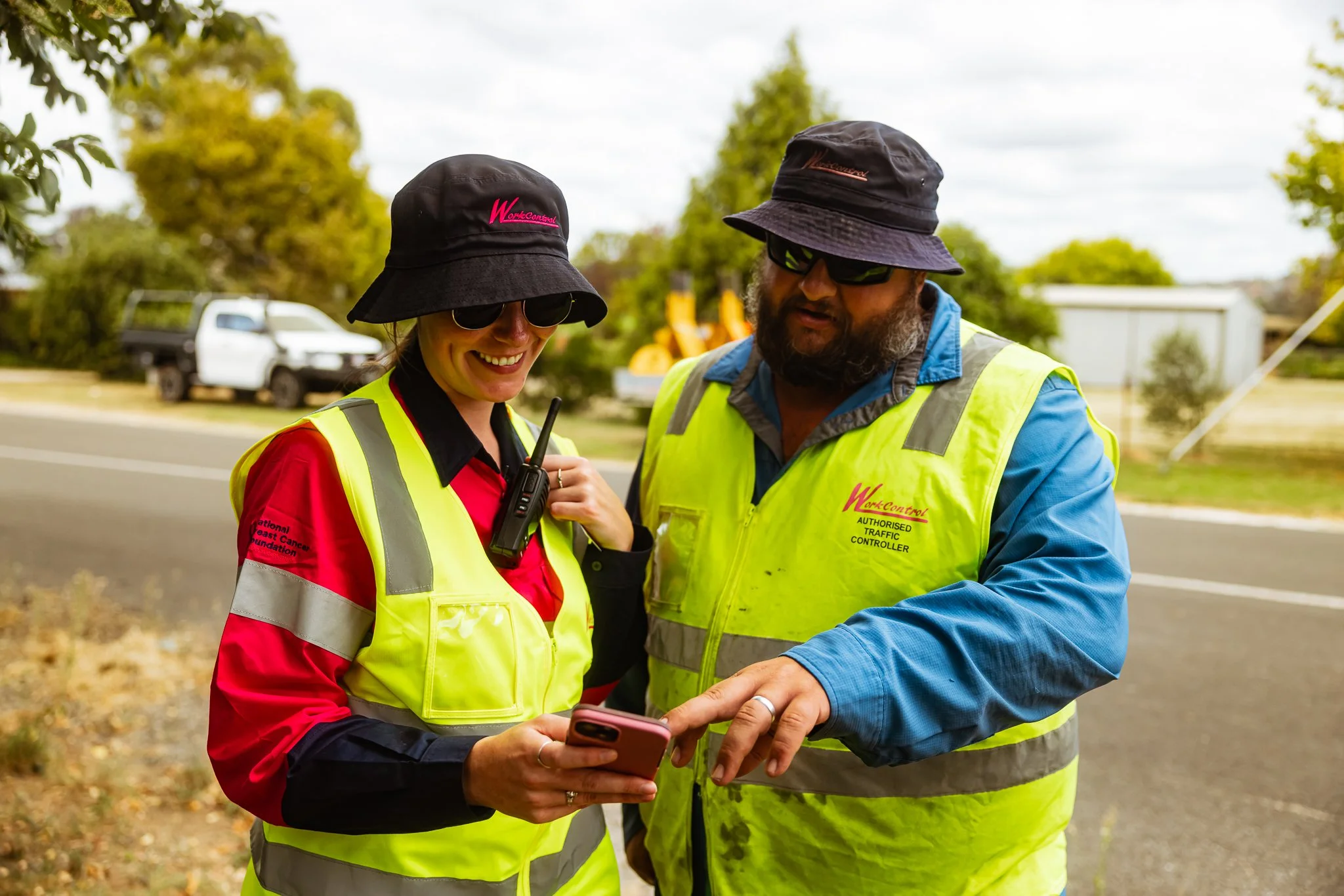 Two traffic controllers in bright yellow safety vests and wide-brim hats smiling and looking at a smartphone together on the roadside. One is holding a walkie-talkie.