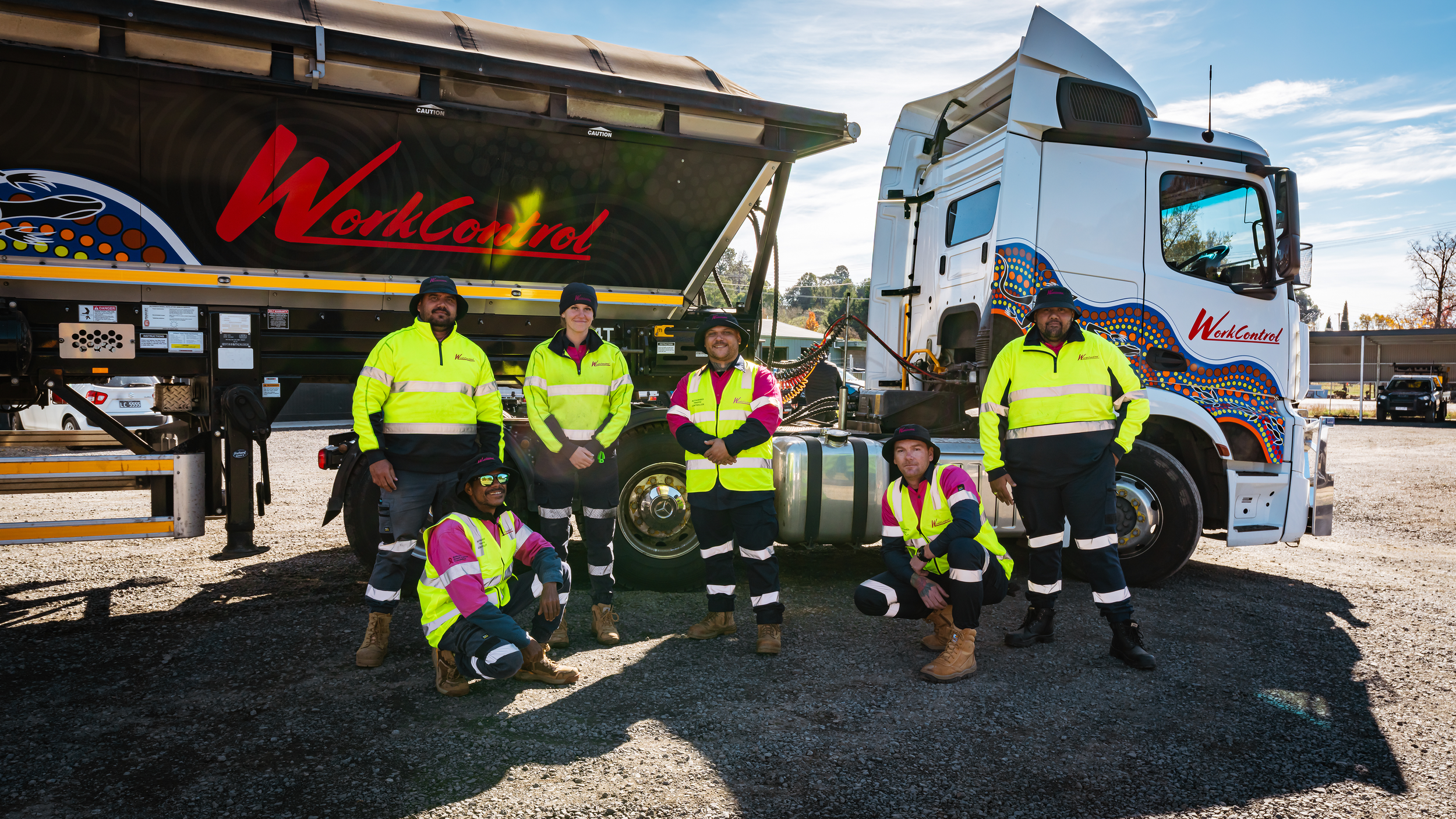 Group of six workers in bright yellow and pink safety jackets posing in front of a large work truck with the logo 'WorkControl' on it.