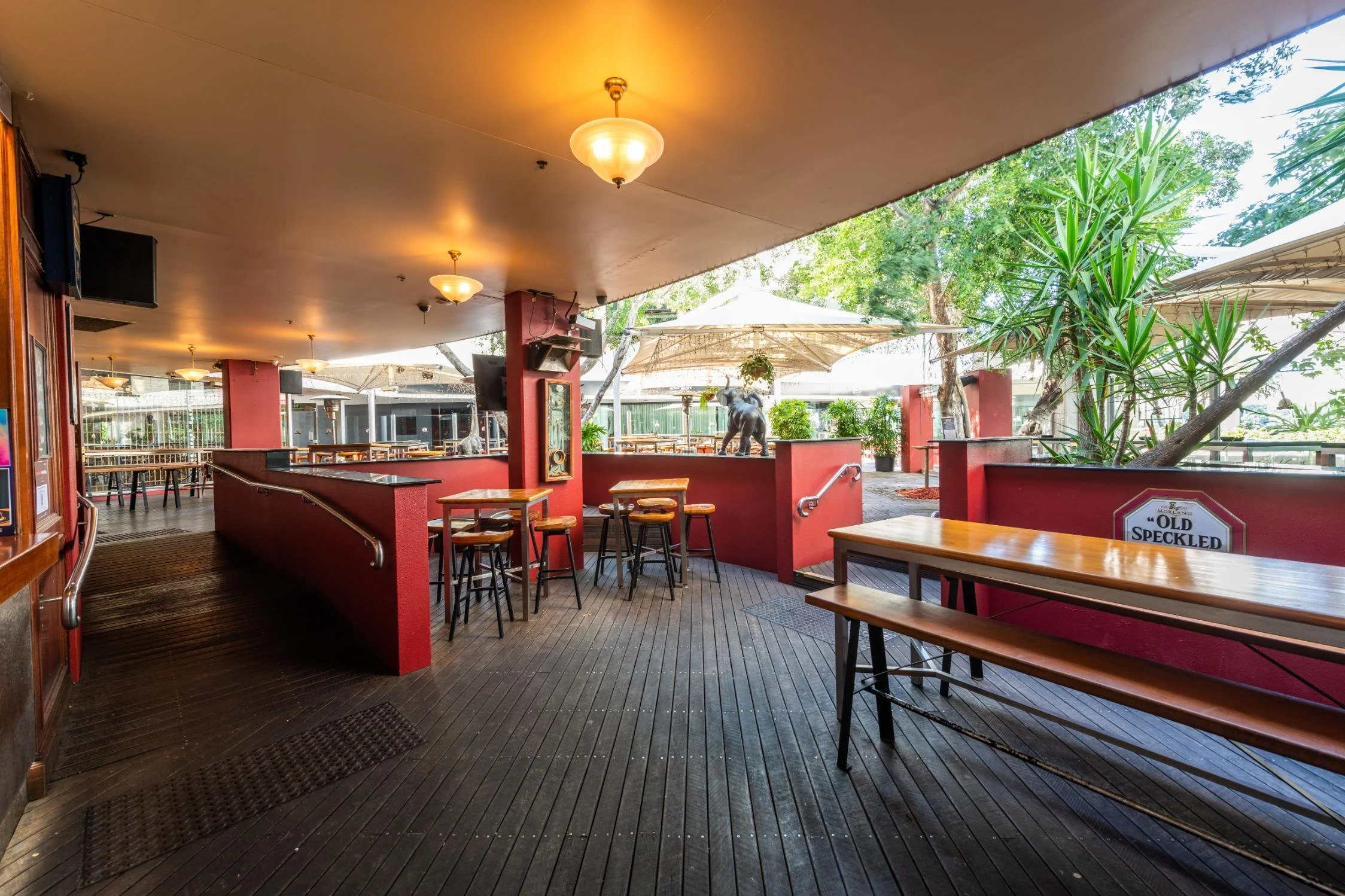 Outdoor patio area of a restaurant with wooden tables, red walls, and potted plants, under large umbrellas, with trees in the background.