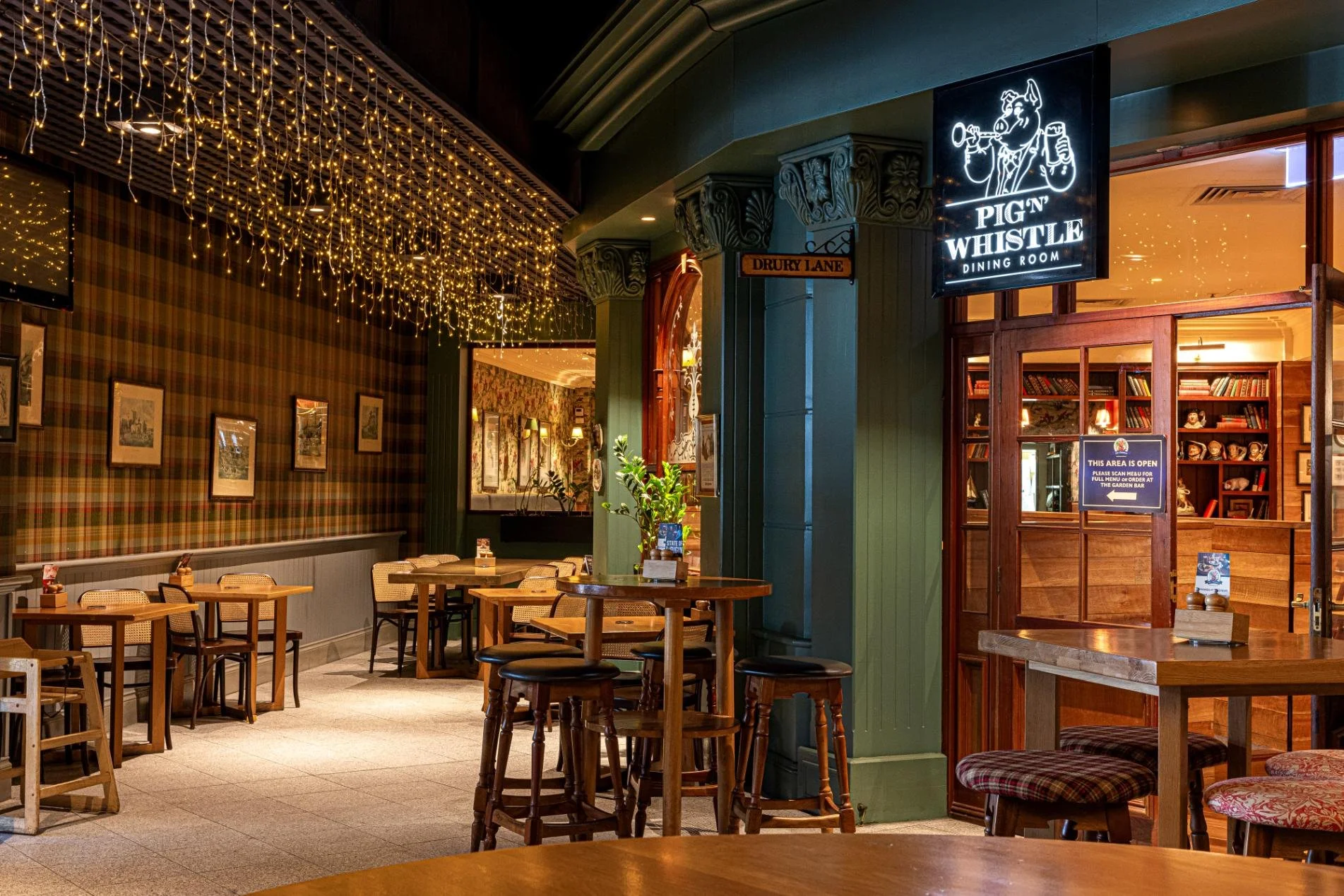 Interior of a cozy restaurant with warm lighting, decorated with string lights hanging from the ceiling, featuring wooden tables and chairs, some with plaid cushions, and a sign for "Pign Whistle Dining Room".