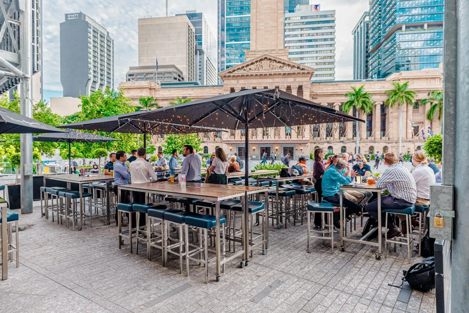 People dining outdoors at a restaurant with tables shaded by black umbrellas, city buildings in the background, and string lights hanging from the umbrellas.