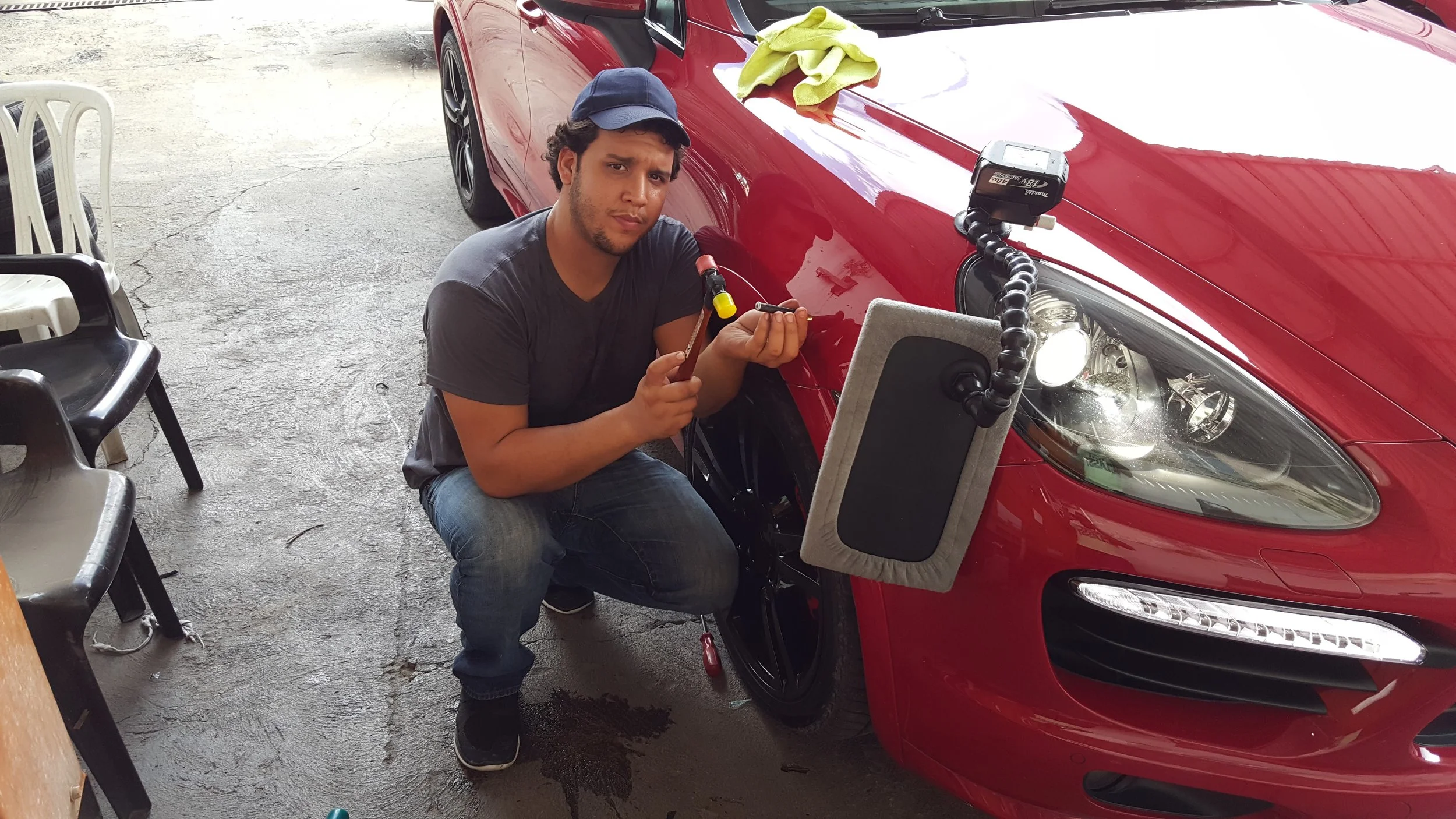 Angel pulling dents on a red Porsche in a garage, crouching beside the front wheel, holding a tool. The car's headlight is visible, and a cleaning cloth is on the car's hood. There are chairs and a concrete floor around.