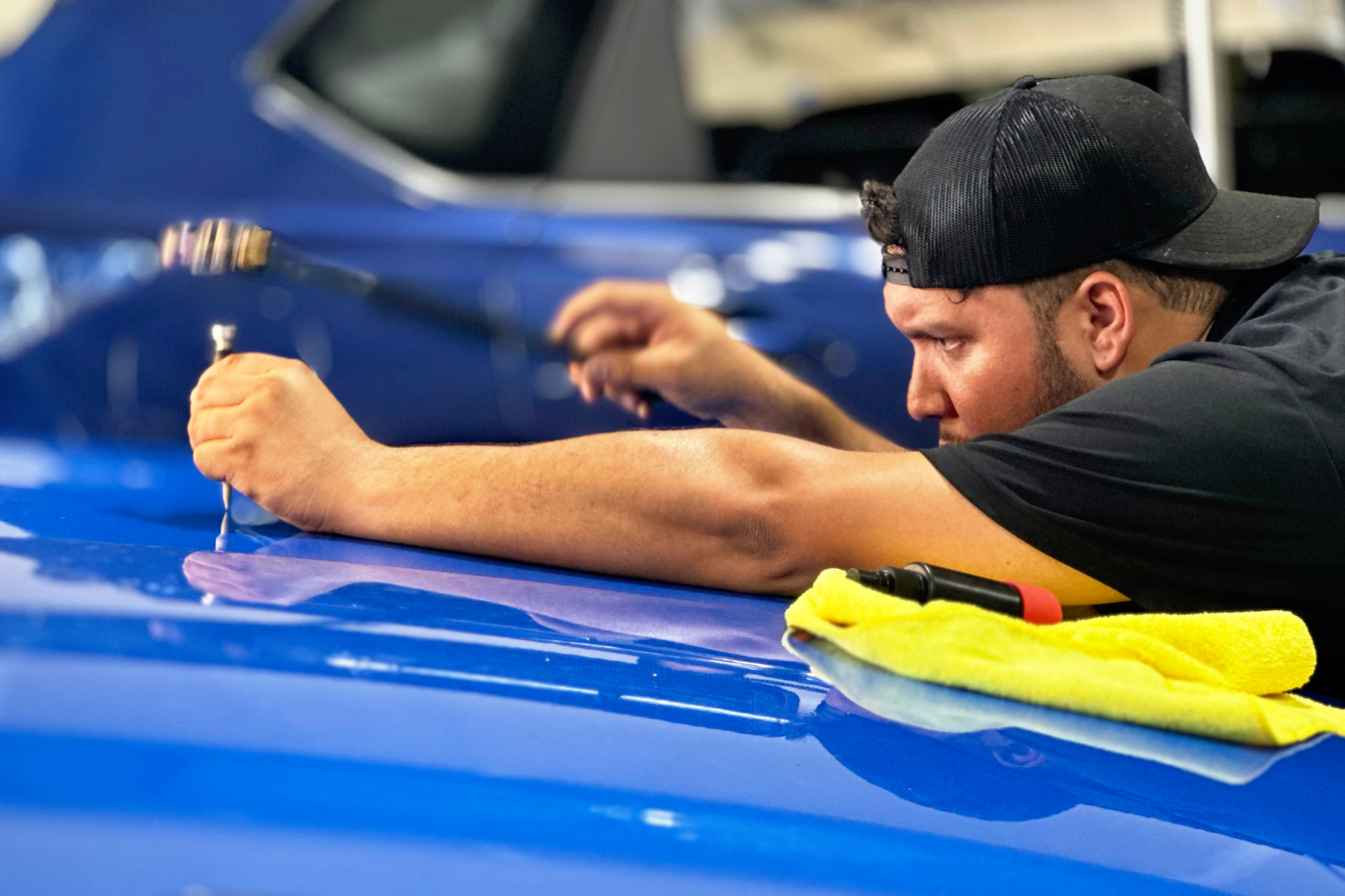 A man wearing a black cap and black shirt is installing or repairing a blue car, leaning on the hood and using a tool. A yellow cloth and a screwdriver are on the car's hood.