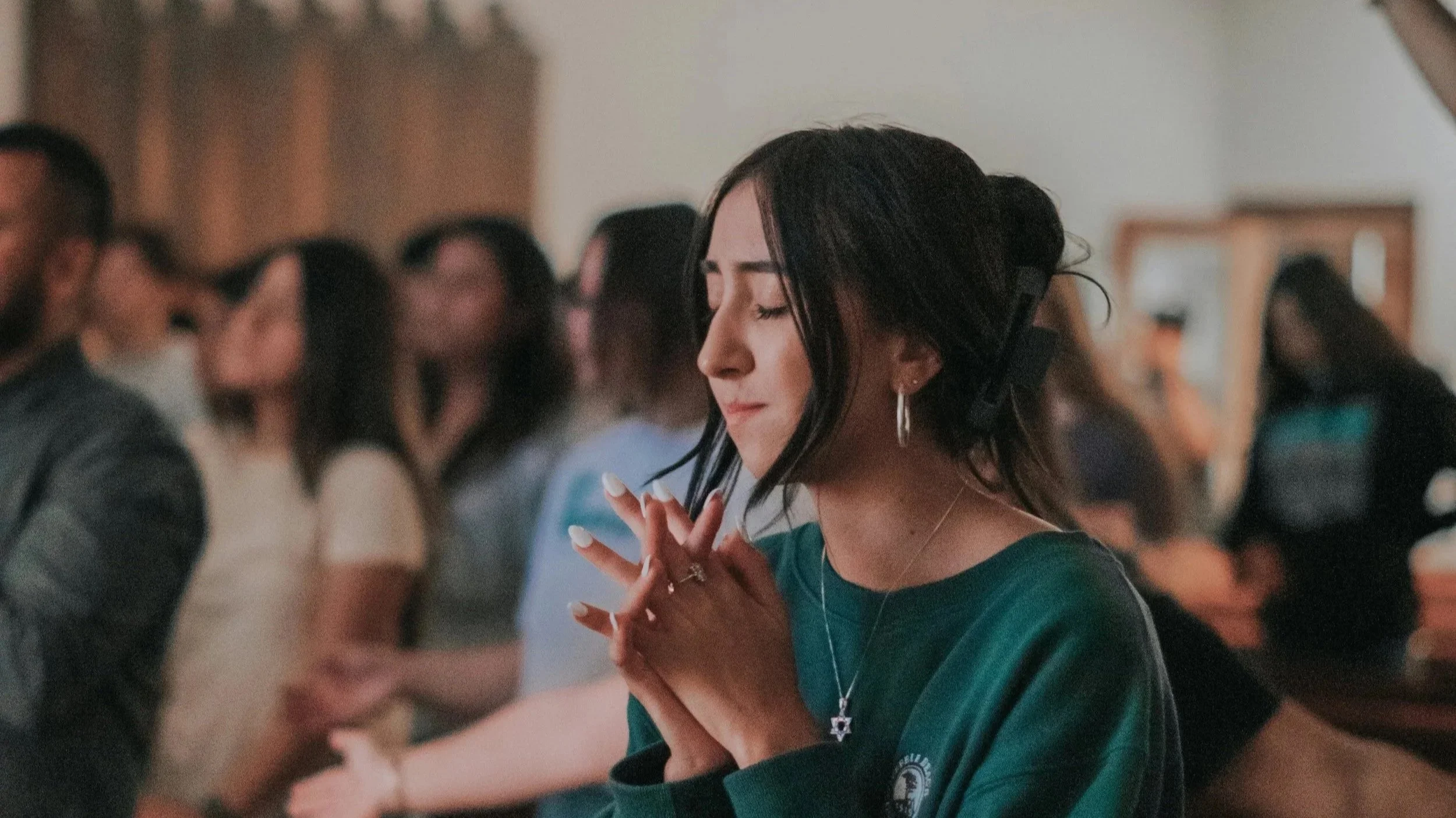 A young woman with dark hair in a bun, wearing a green shirt, earrings, and a necklace, has her eyes closed and hands clasped in prayer or reflection at a gathering.
