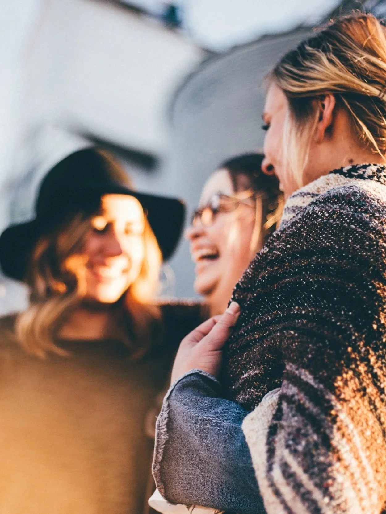 Three women laughing and smiling, one wearing a wide-brimmed hat, another with glasses, and the third with blonde hair, close together outdoors under sunlight.